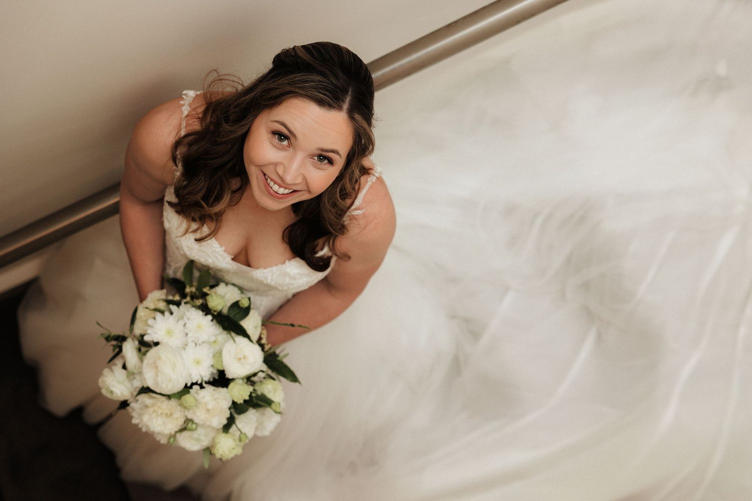 Overhead view of bride in white dress holding elegant white rose bouquet while sitting on stairs.