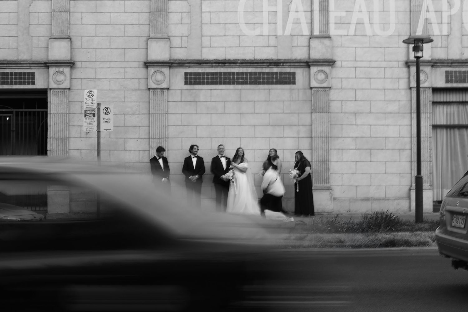 A black and white wedding party stands outside a building as a car blurs past in the foreground.