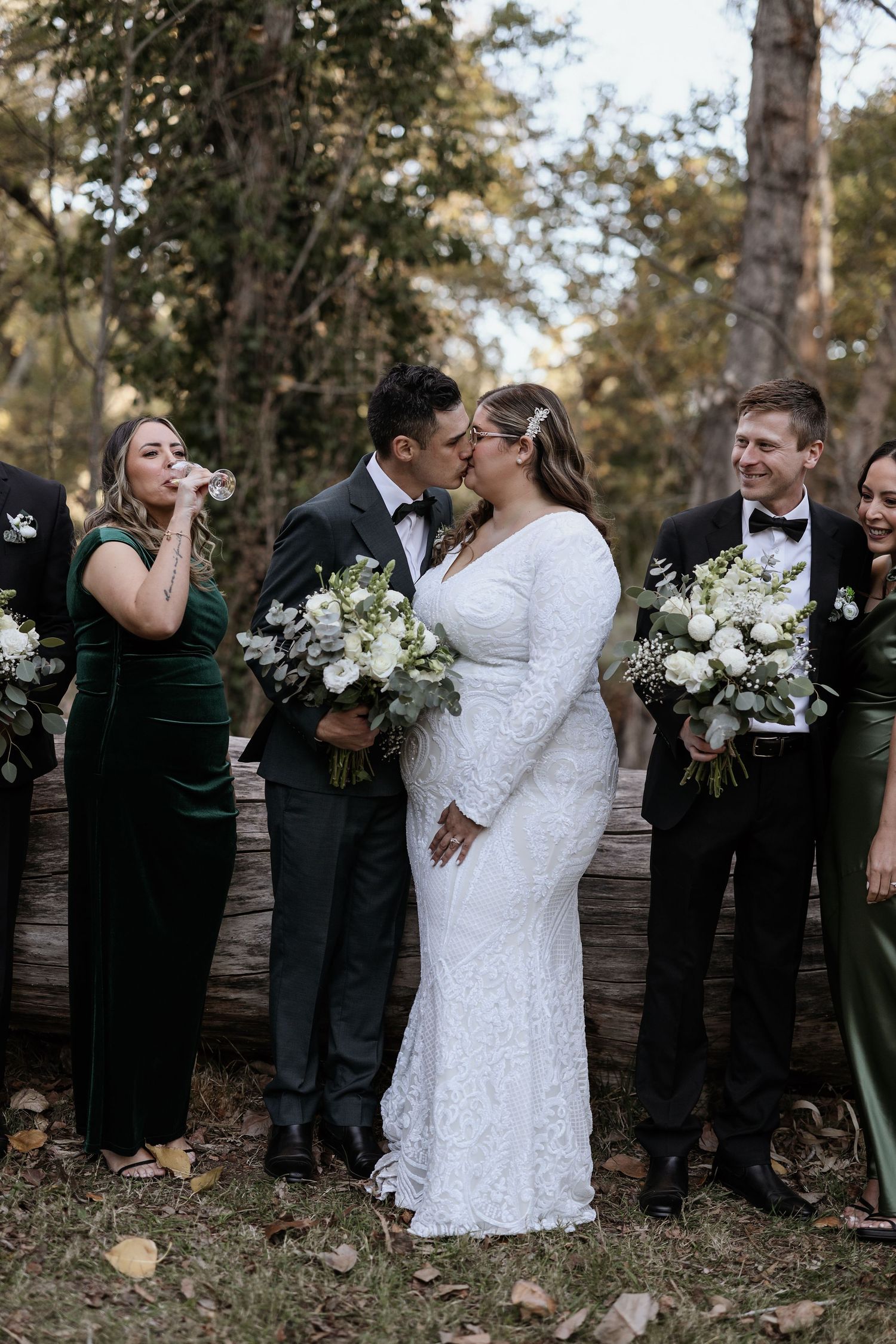 Wedding party in emerald green dresses and black tuxedos gather outdoors for photos with white floral bouquets.