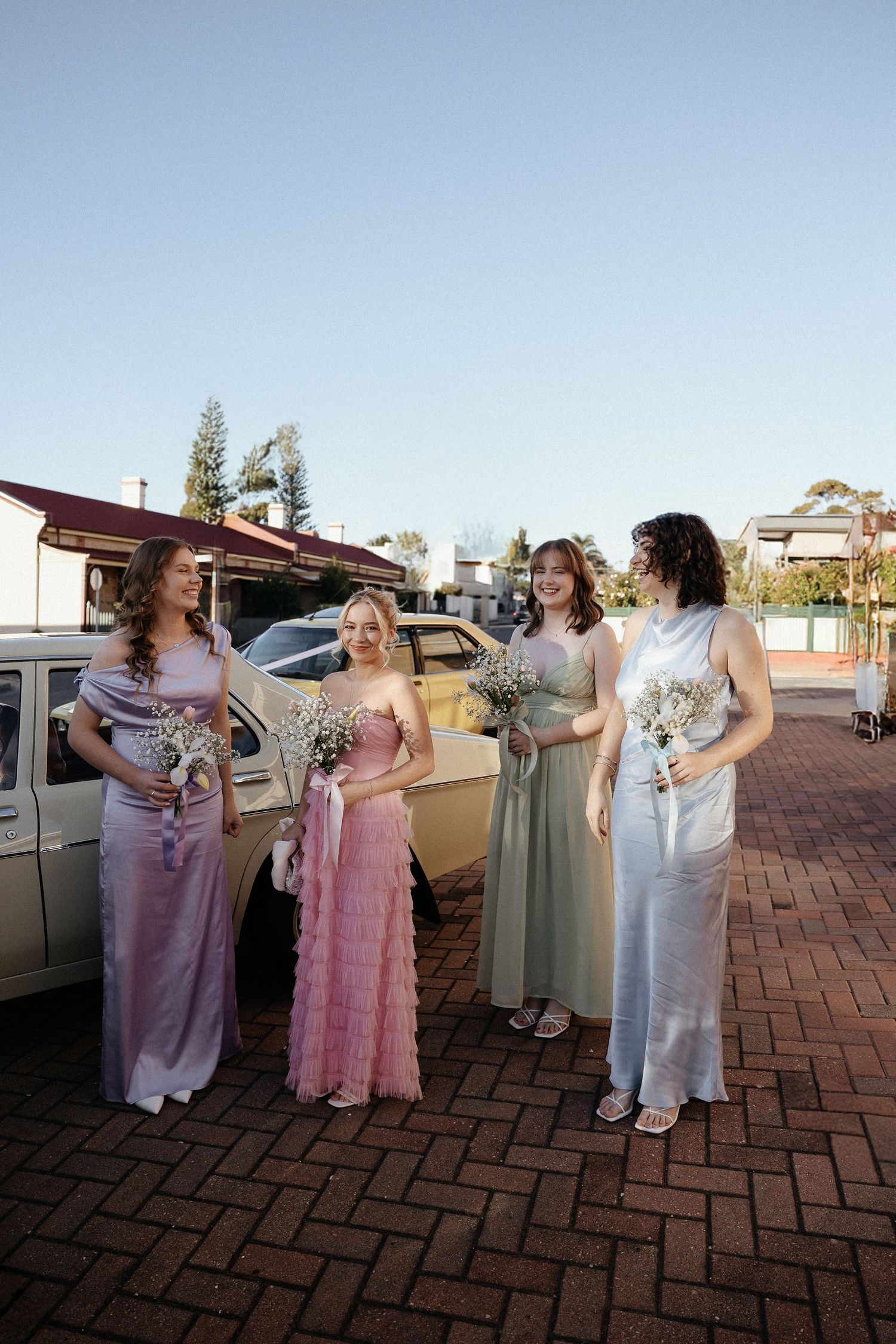 Bridesmaids in elegant floor-length dresses stand together on brick pavement next to a vintage car on a sunny day.