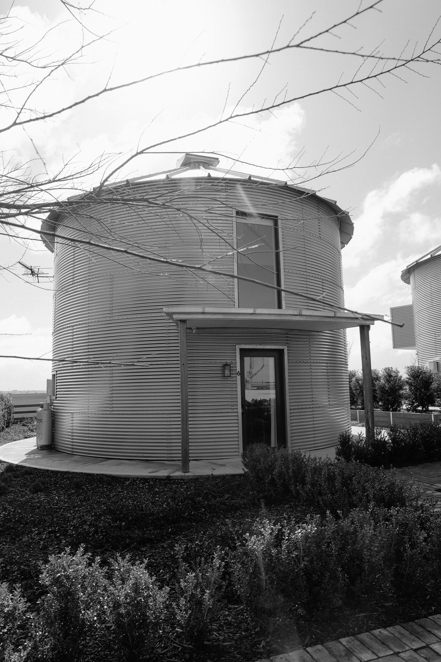 Large industrial grain storage silos stand in a row against a cloudy sky with bushes growing in the foreground.