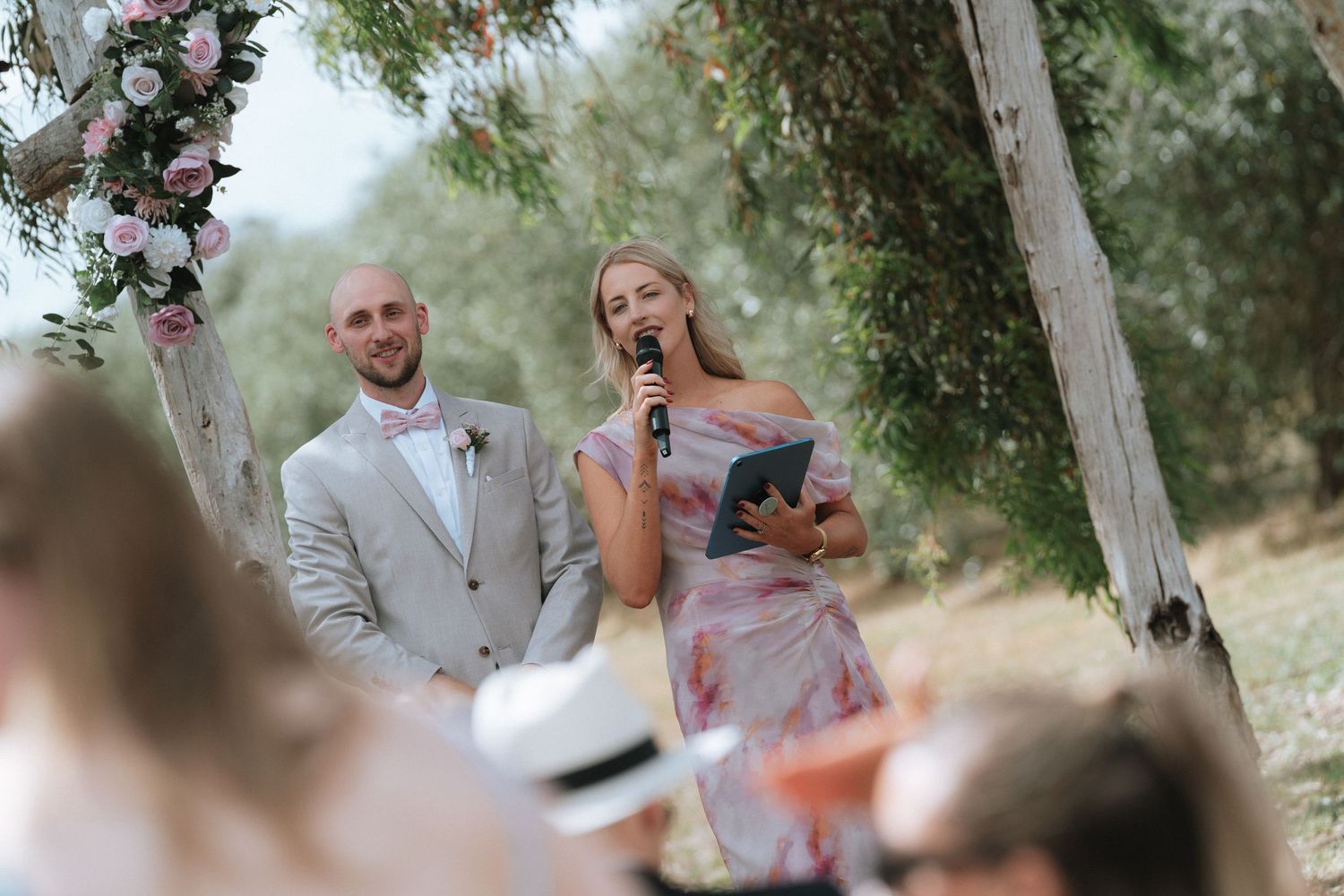 A romantic outdoor wedding ceremony takes place under a floral arch in a rustic garden setting.
