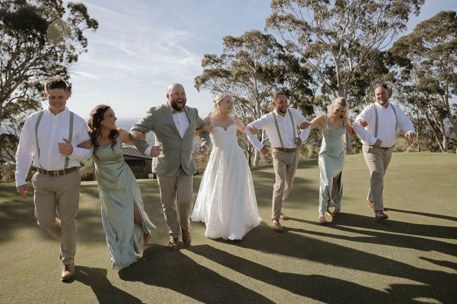 Wedding group forms a fun diagonal line across a sunny outdoor field with trees in the distance.