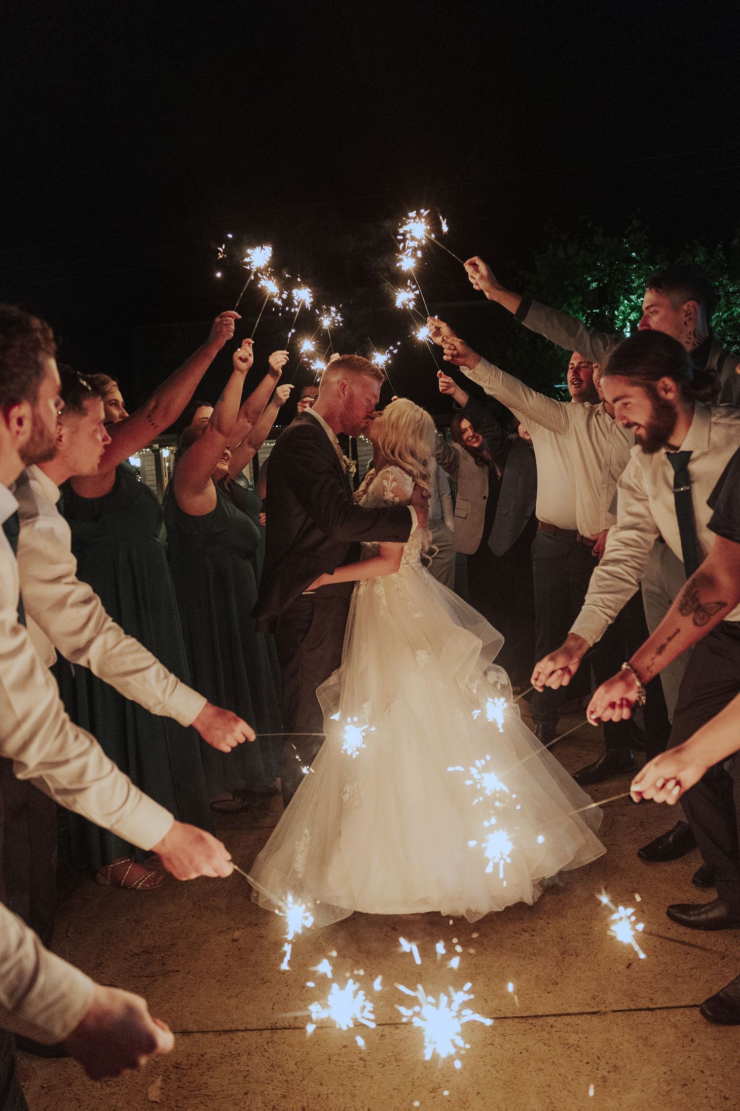 Wedding guests wave sparklers in a circle around a bride in a white ballgown during an outdoor nighttime celebration.