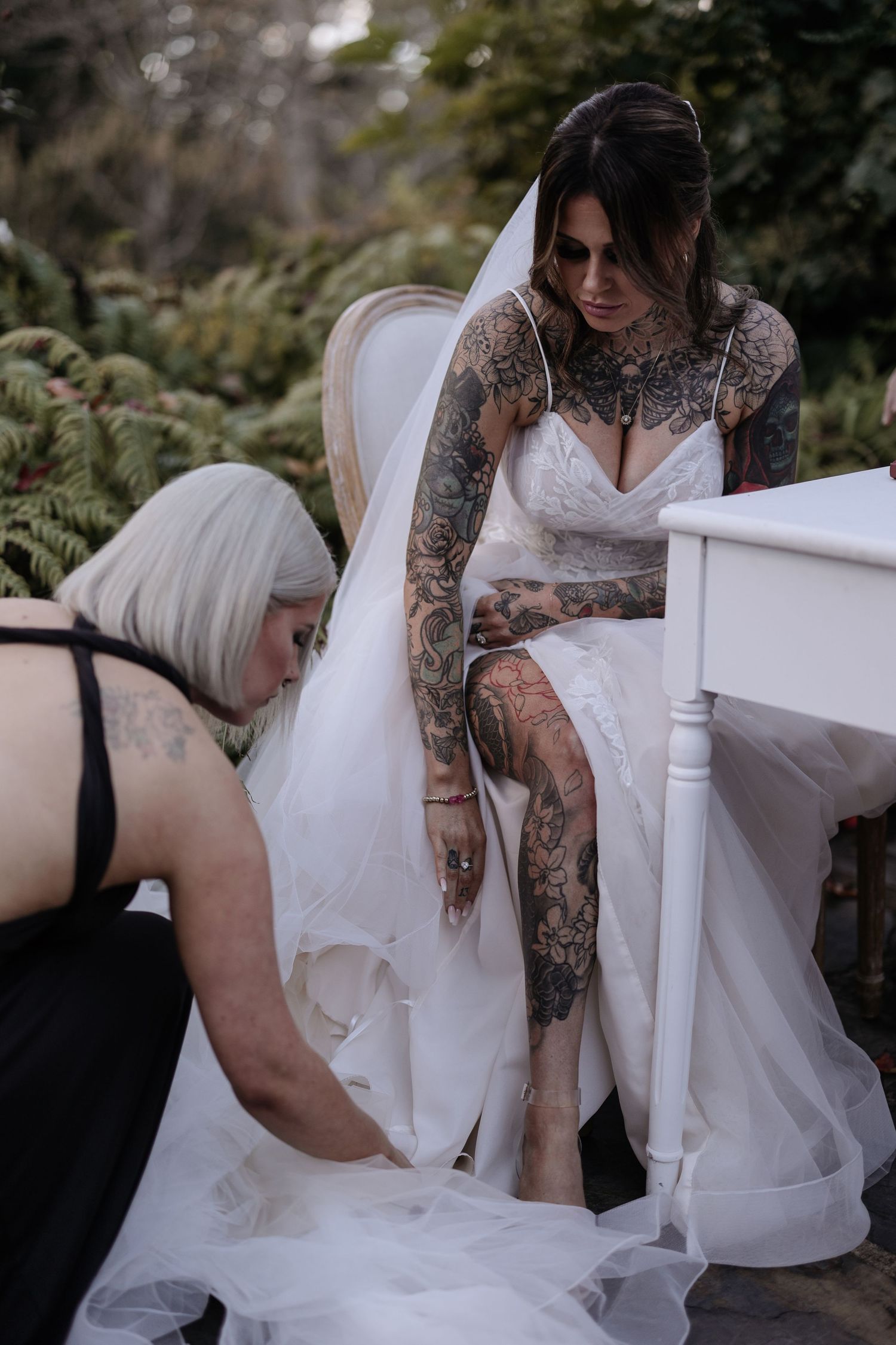 A bride in a white wedding dress with intricate tattoos gets help with her gown from someone adjusting the hem.