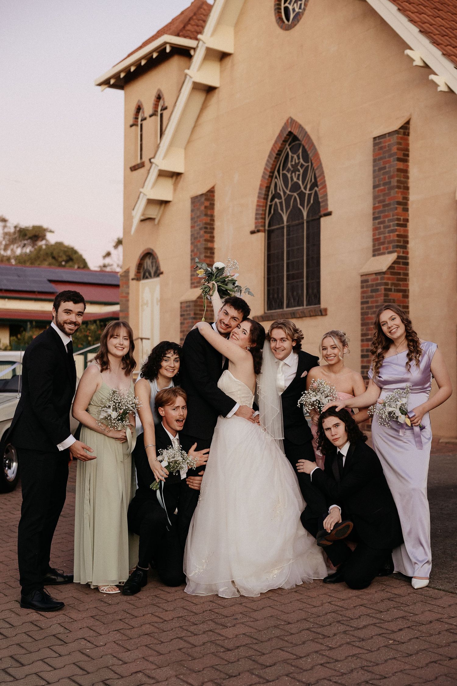 Wedding party poses playfully together outside a church with brick and stucco exterior in warm evening light.