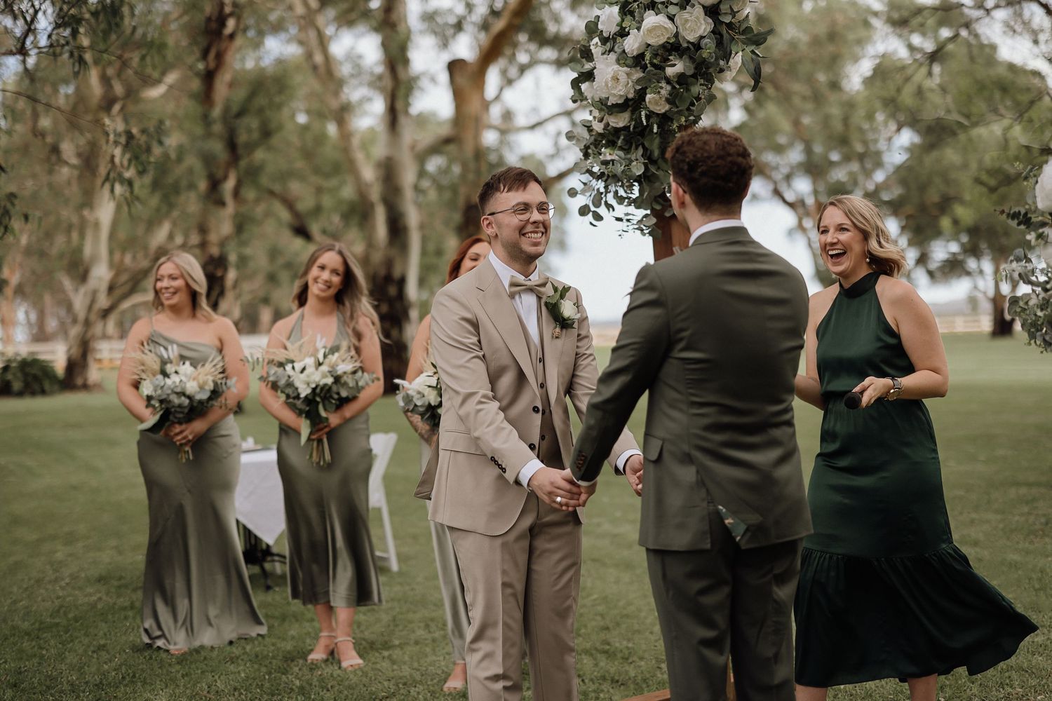 Wedding party celebrates joyfully on a grassy lawn with trees in the background.