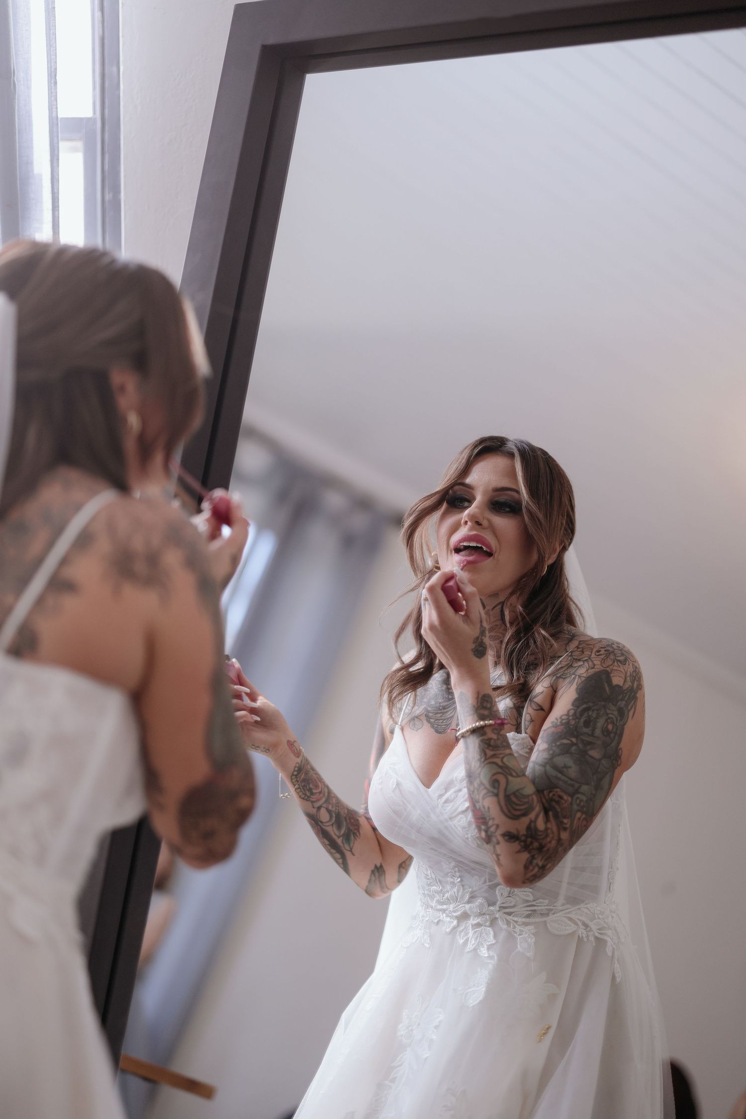 A person in a white dress applies makeup while getting ready in a bright room with natural lighting.