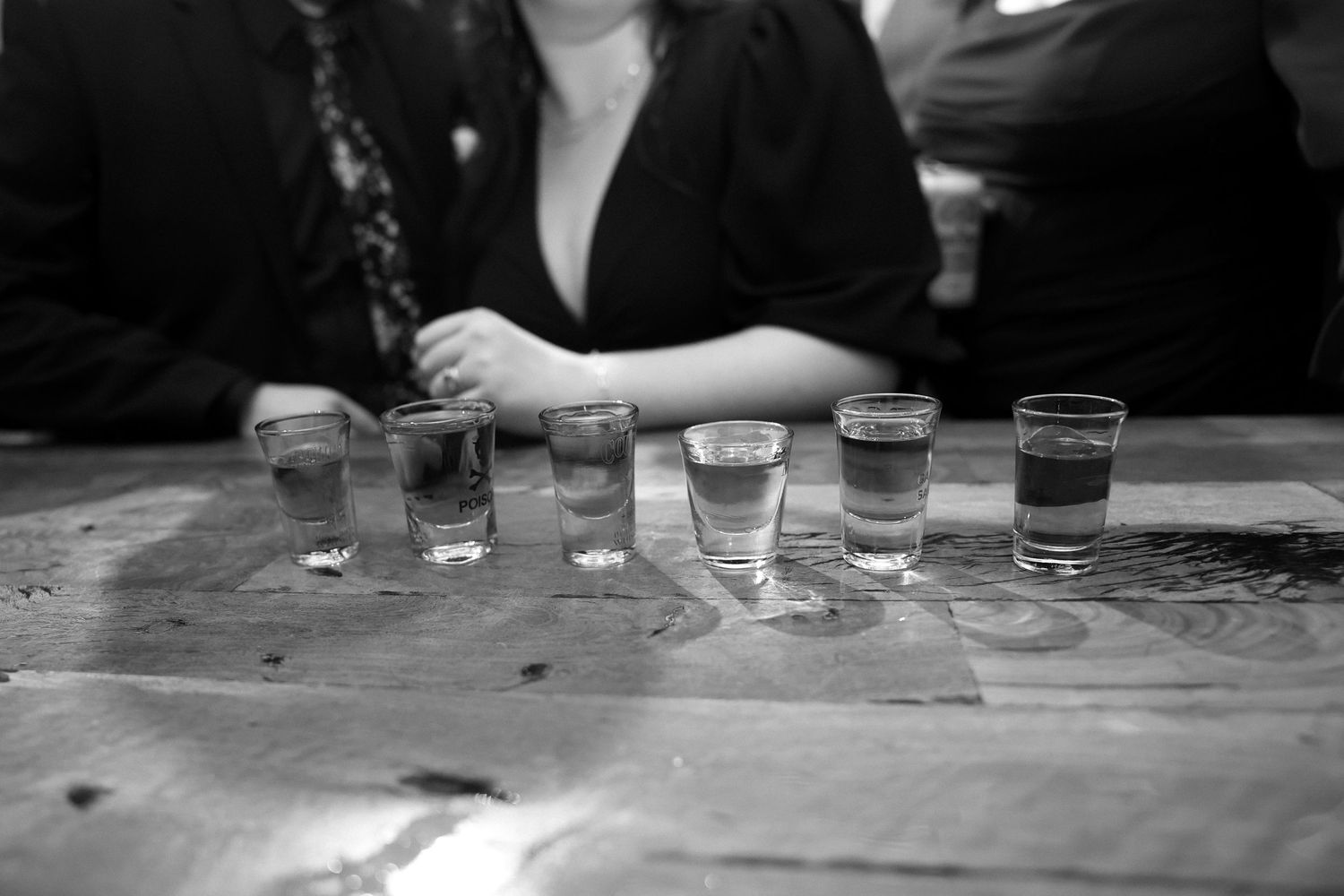A black and white photograph shows a row of empty shot glasses lined up on a wooden surface.