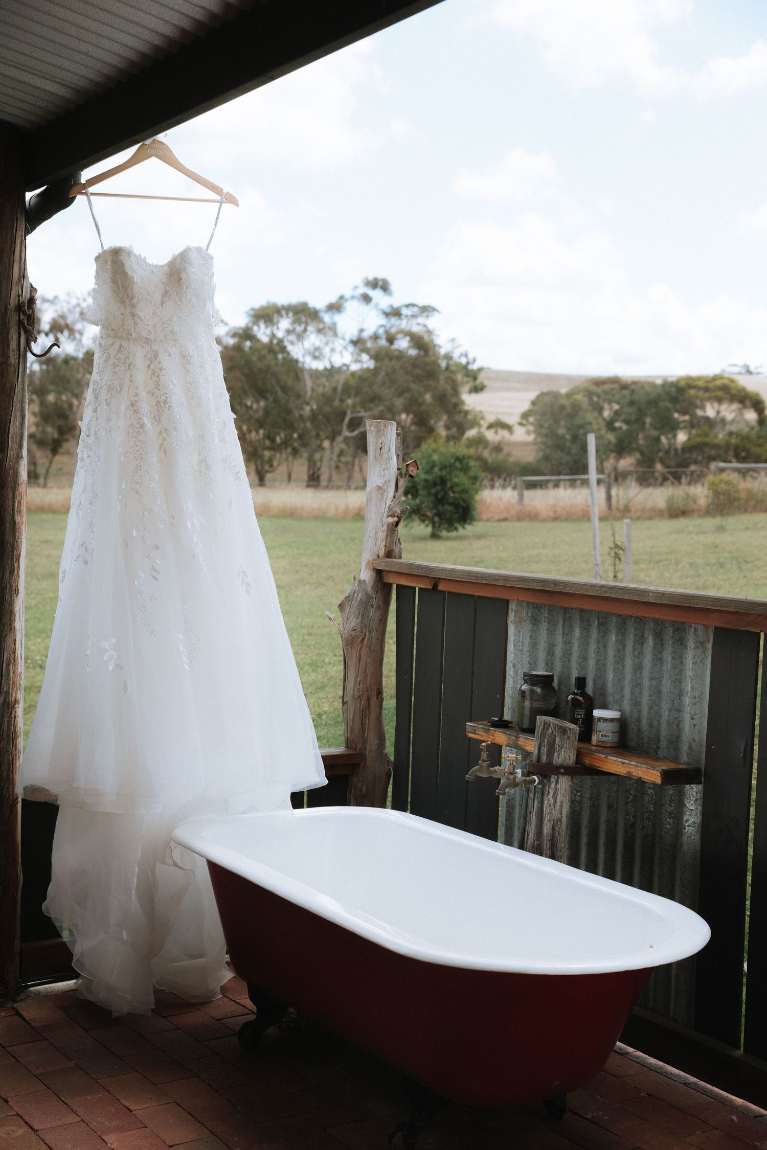 A white wedding dress hangs elegantly against a rustic backdrop with a vintage bathtub and scenic countryside views.