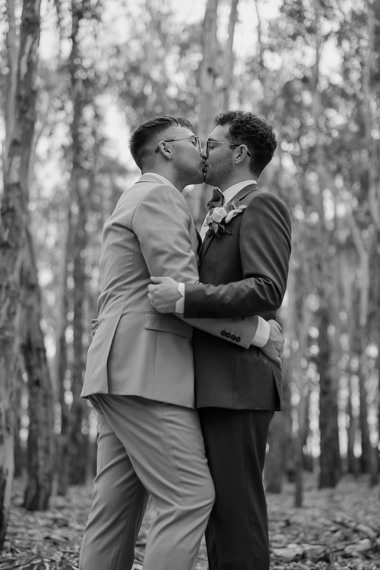 A couple in formal attire shares an intimate moment while embracing in a wooded forest setting.