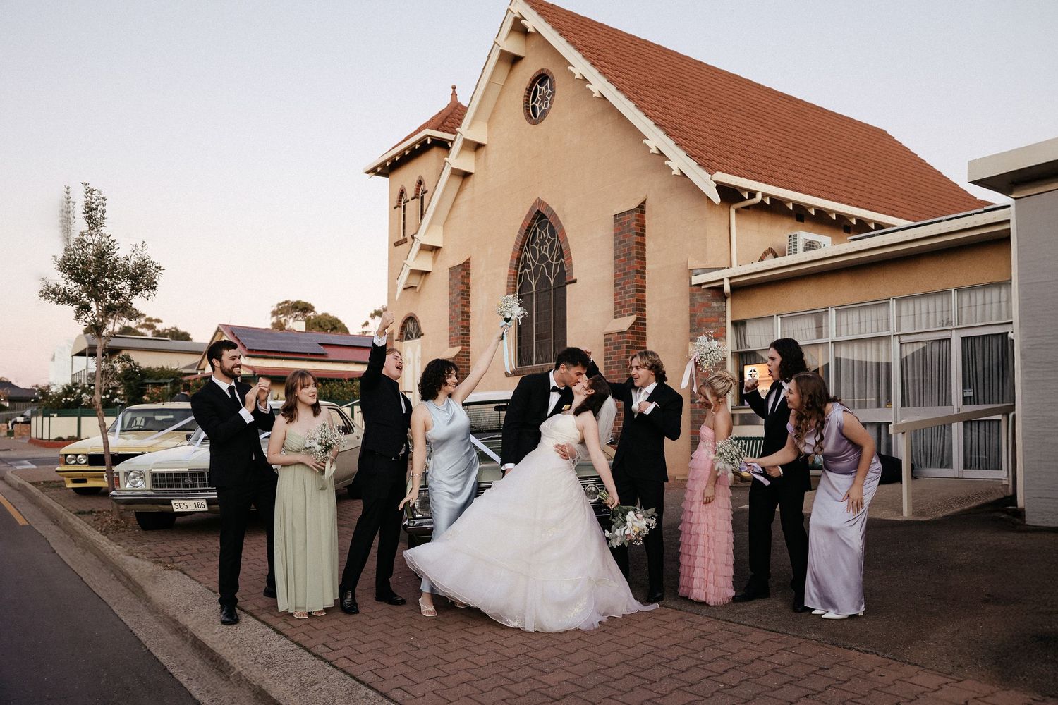 Wedding party in formal attire performs coordinated dance moves on an outdoor patio during golden hour.
