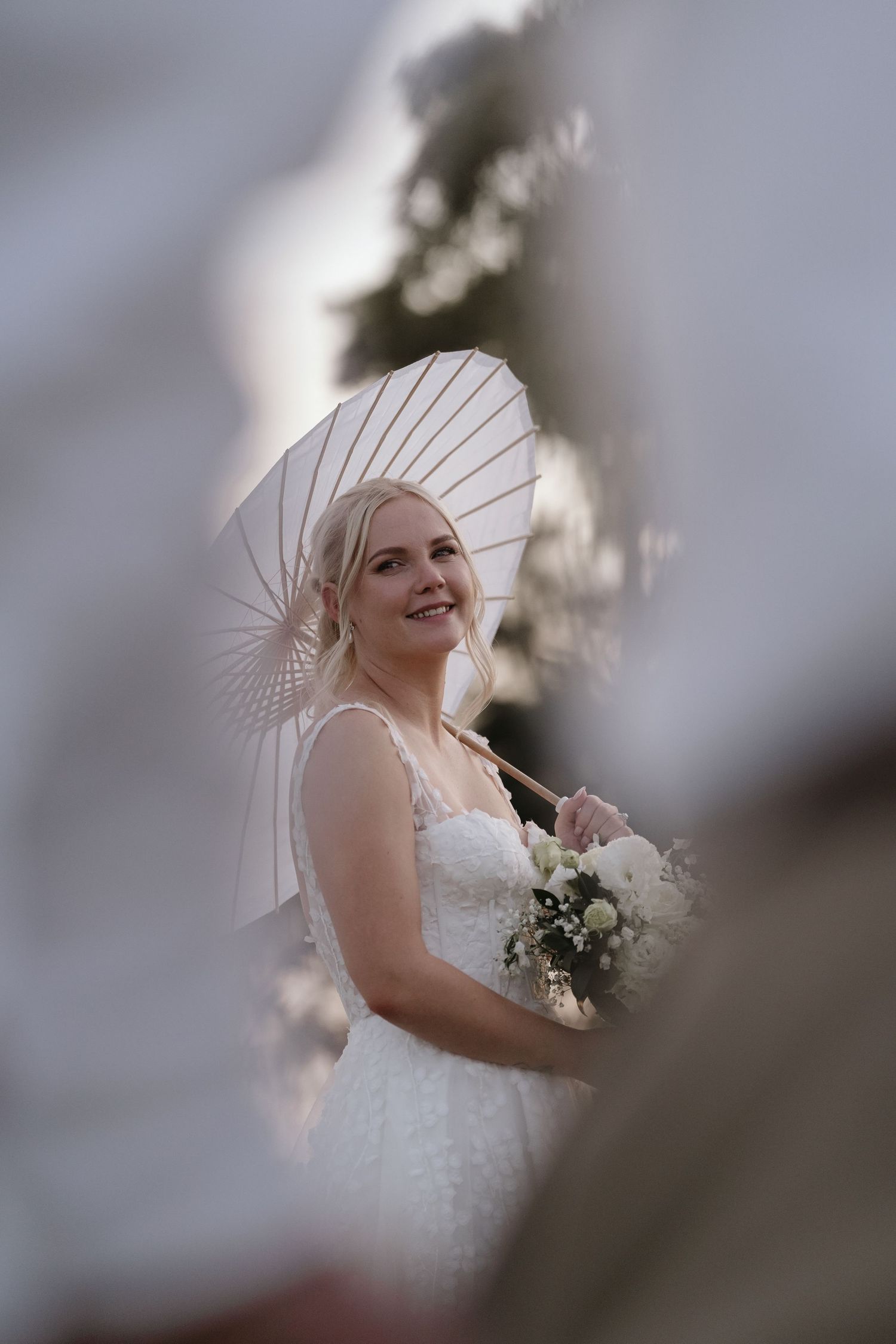 A bride in a white lace dress smiles joyfully while holding a delicate bouquet of flowers during an outdoor wedding.