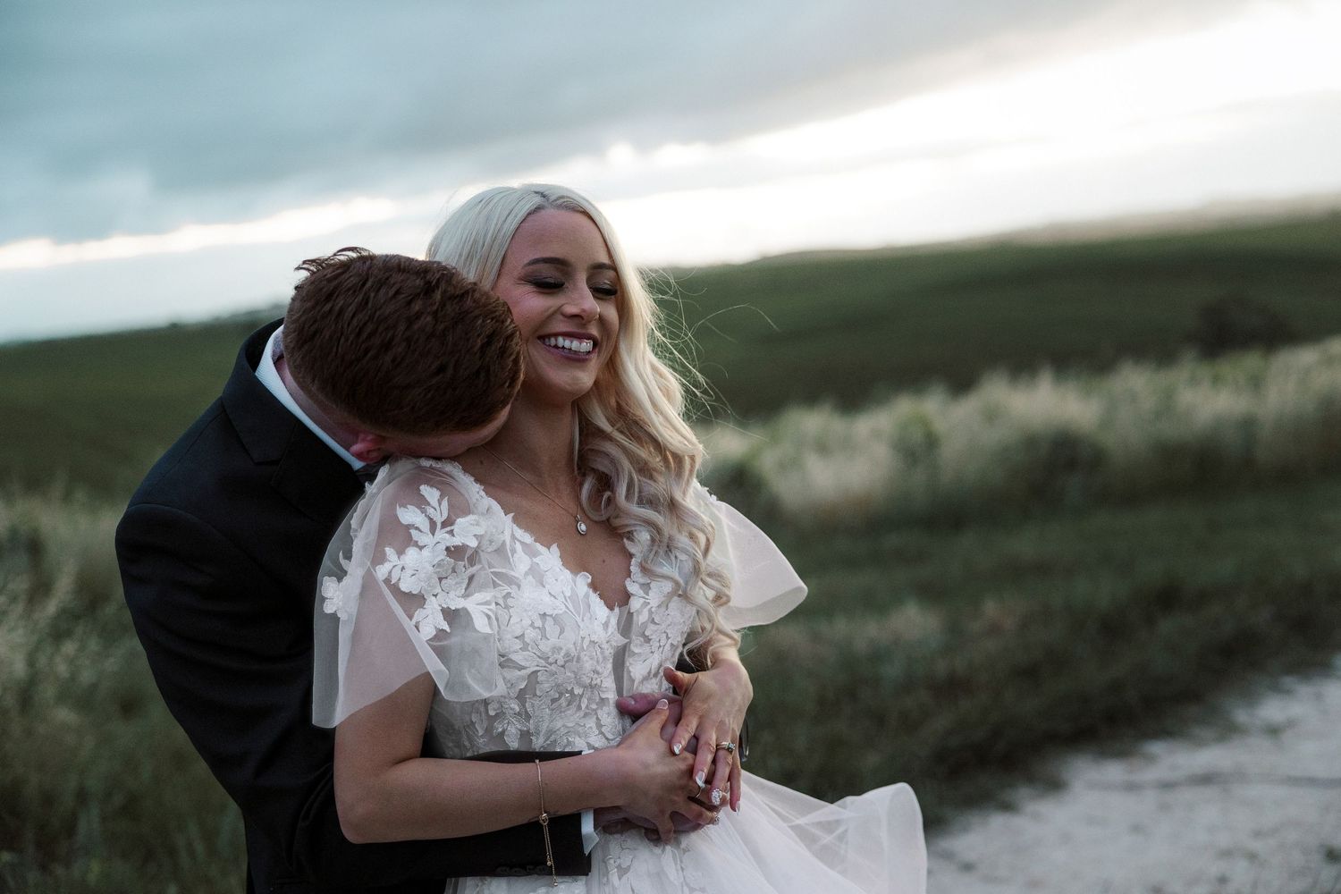 A couple in wedding attire embraces in a scenic field at dusk with dramatic cloudy skies in the background. Gomersal Wines