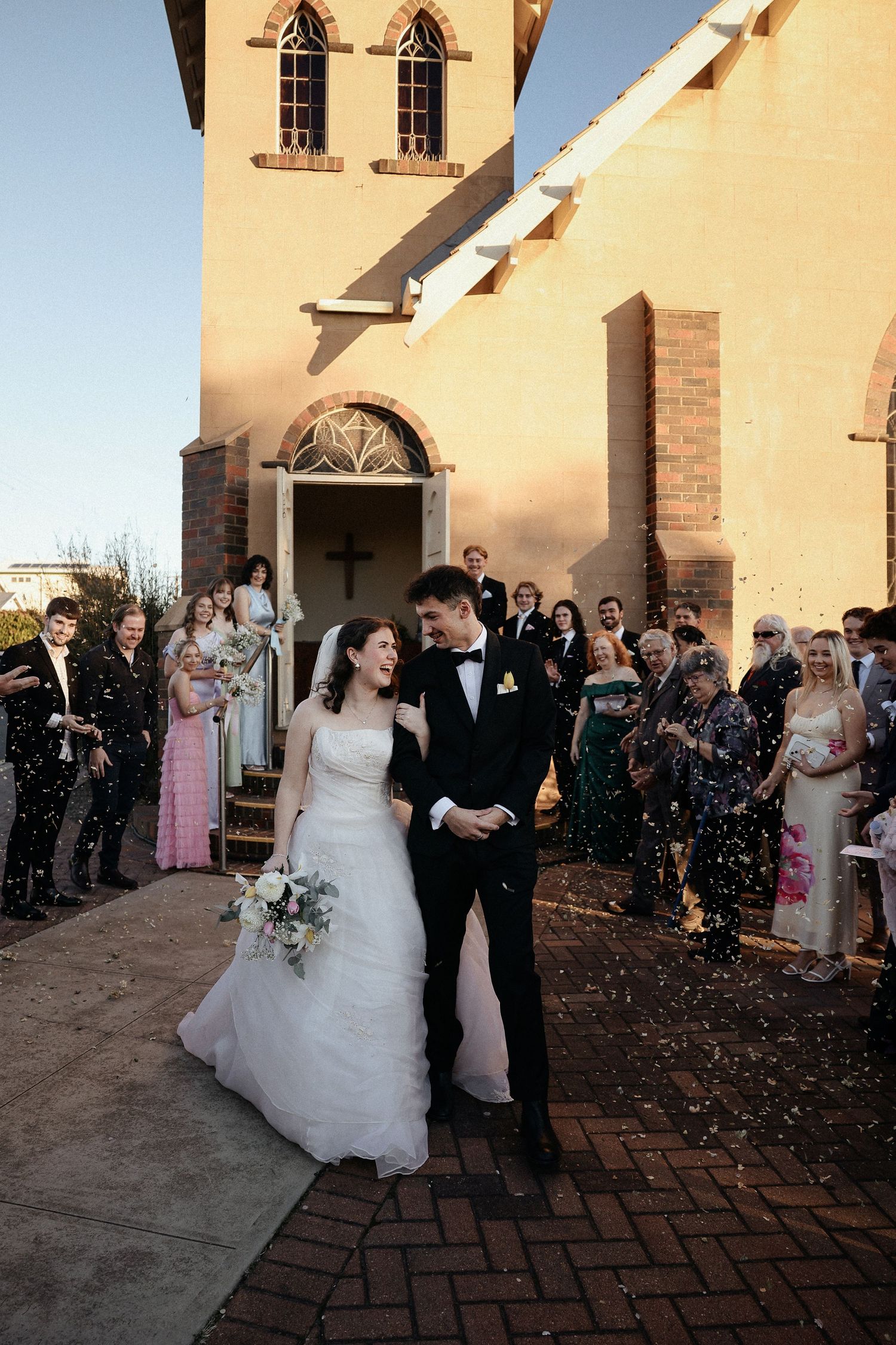 Wedding party exits church while guests throw confetti and celebrate on brick pathway outside historic building.