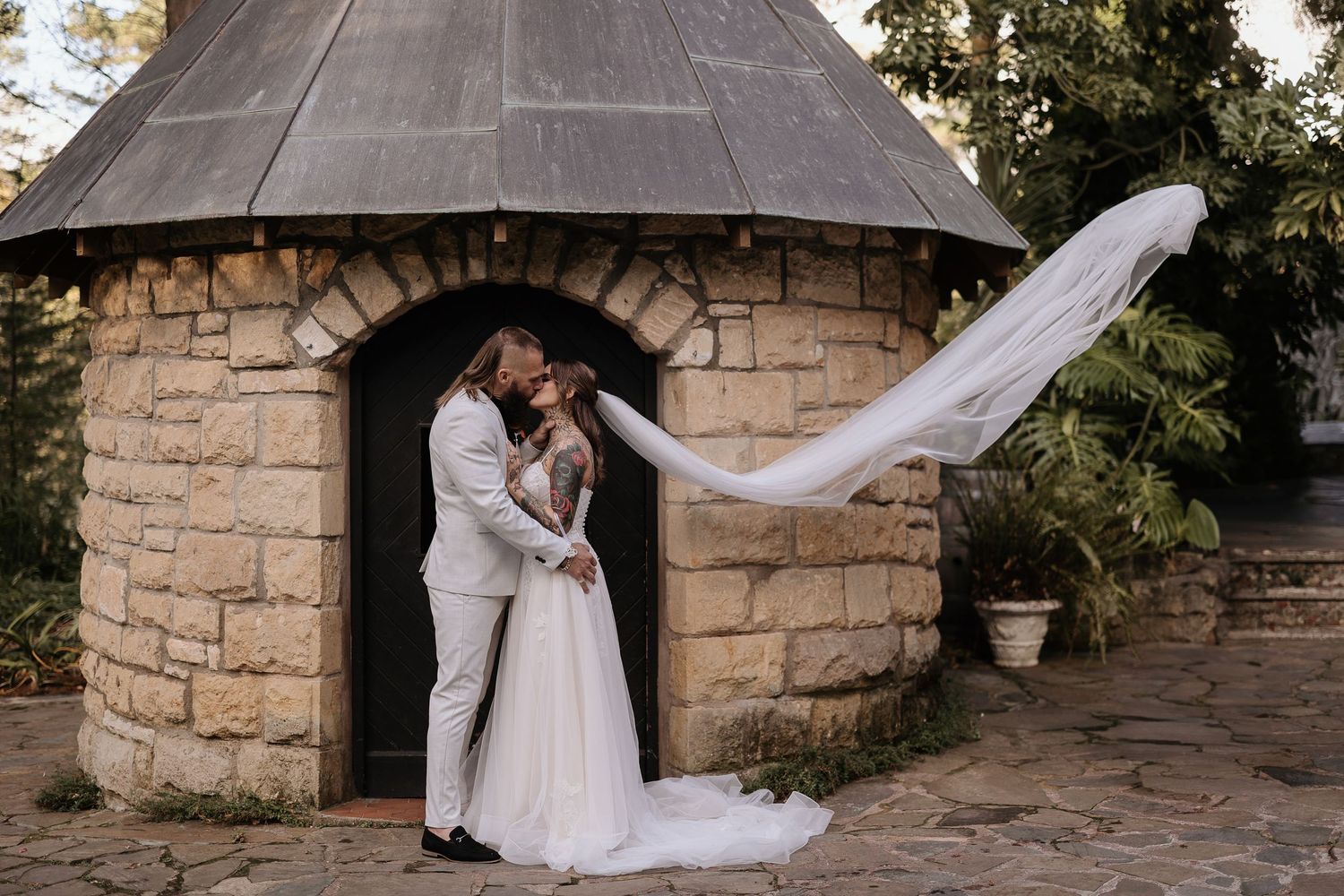 A romantic wedding couple embraces near a stone wall as a flowing veil catches the breeze in an outdoor setting.