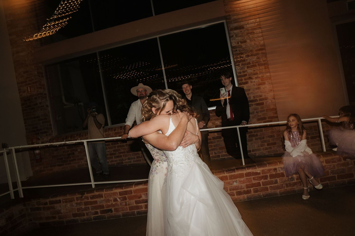 People dancing and celebrating at a reception in a rustic brick-walled venue.