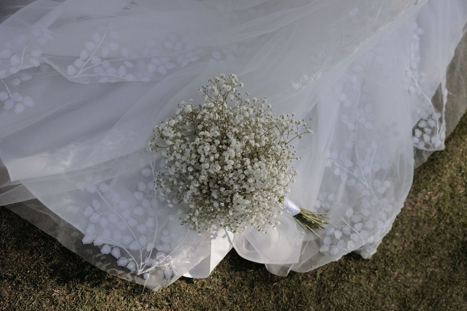 A white wedding dress train with baby's breath flowers lies on grass creating a romantic bridal detail.