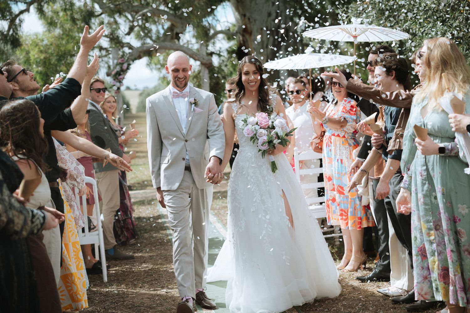 Wedding guests form a celebratory tunnel under white umbrellas for the newlyweds in an outdoor garden setting.