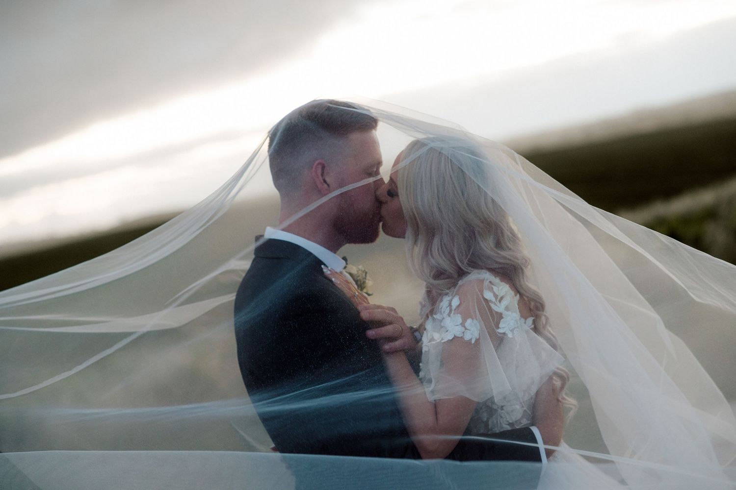 A romantic wedding couple shares an intimate kiss under a flowing white veil in an outdoor setting. wedding photo ideas