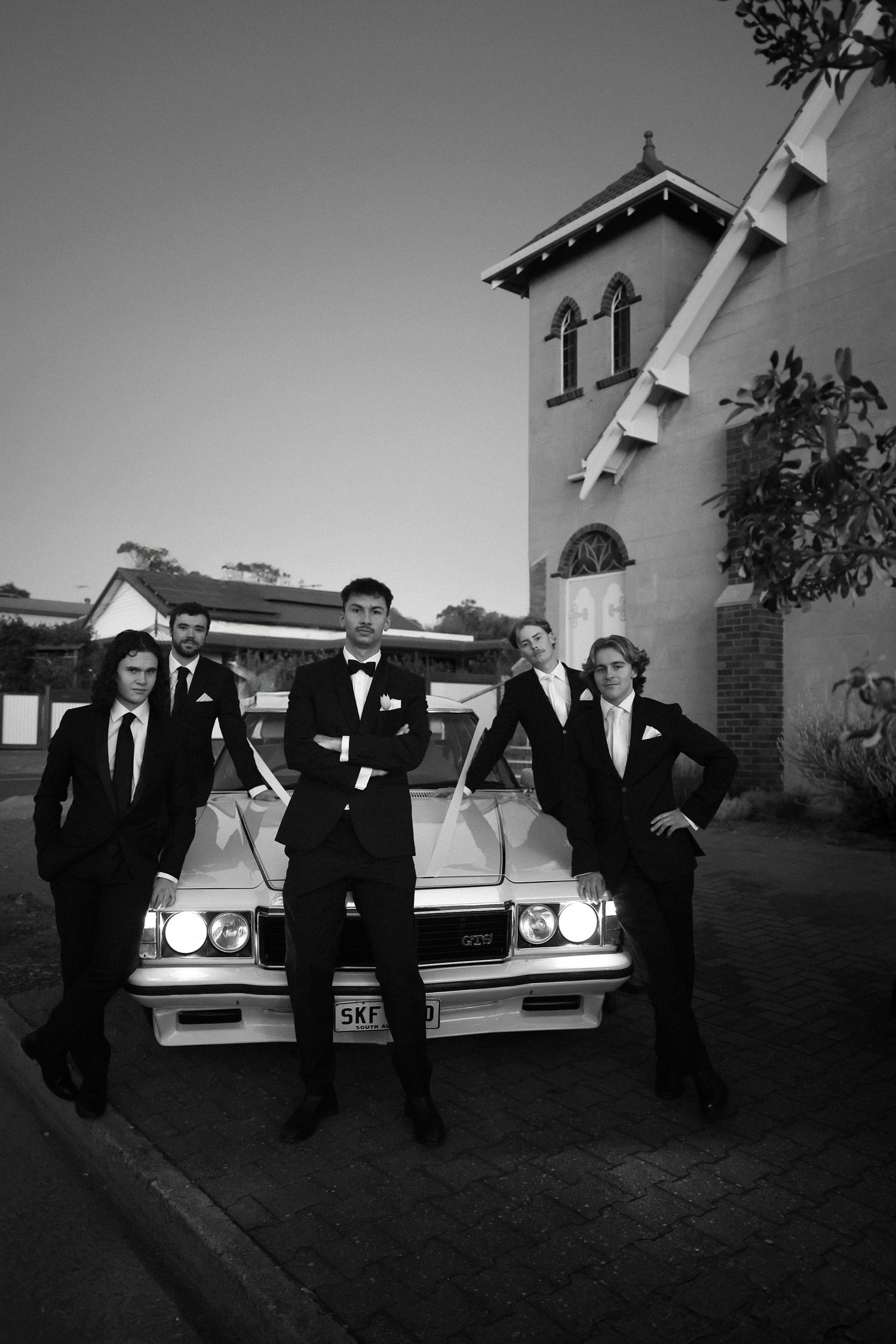 Groomsmen in formal attire pose with a classic car outside a church building.