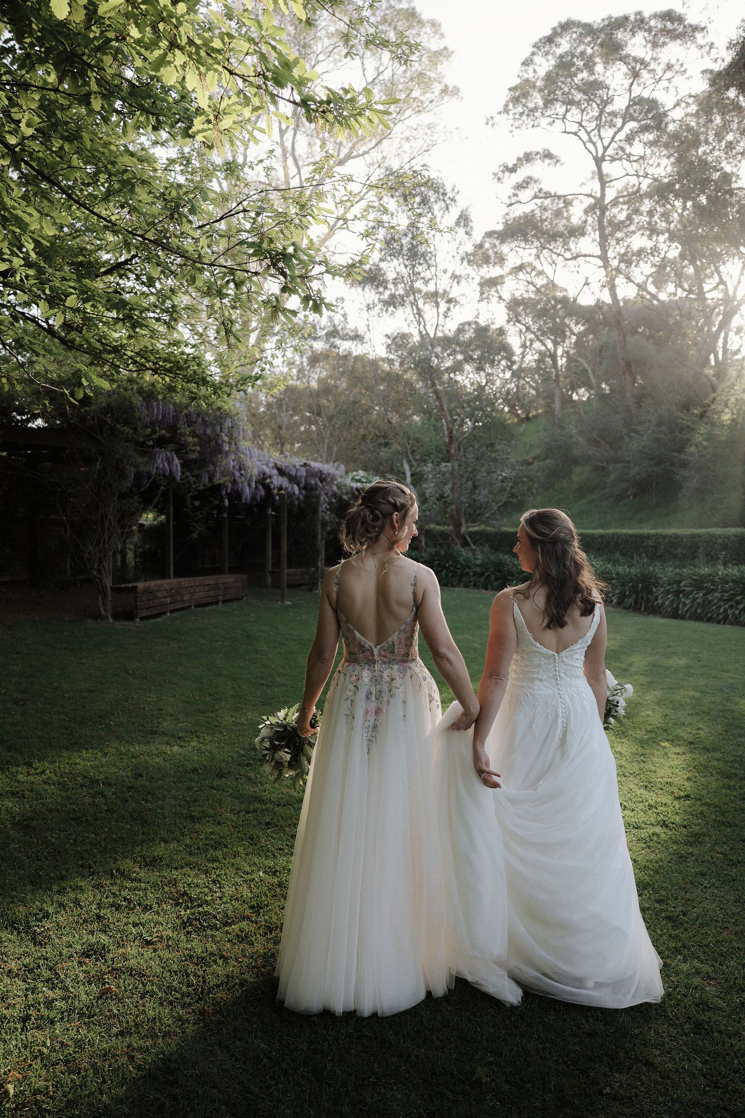 Two people in white wedding dresses sit together on grass at sunset in a garden setting.