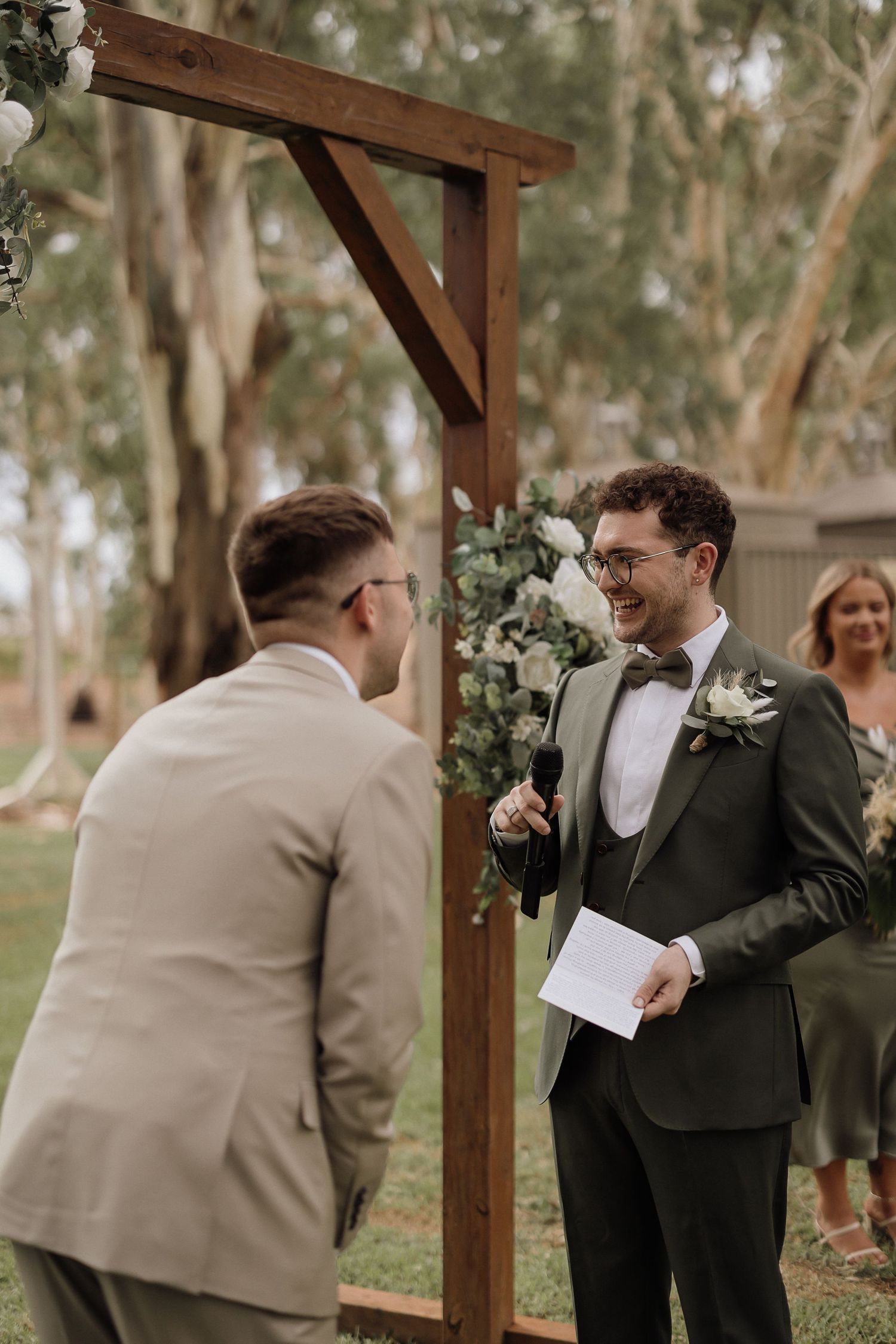 Wedding ceremony taking place under a wooden pergola surrounded by eucalyptus trees during an outdoor celebration.