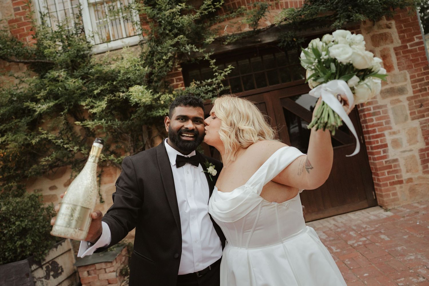 A couple shares a romantic moment in front of a rustic brick wall during a wedding celebration.