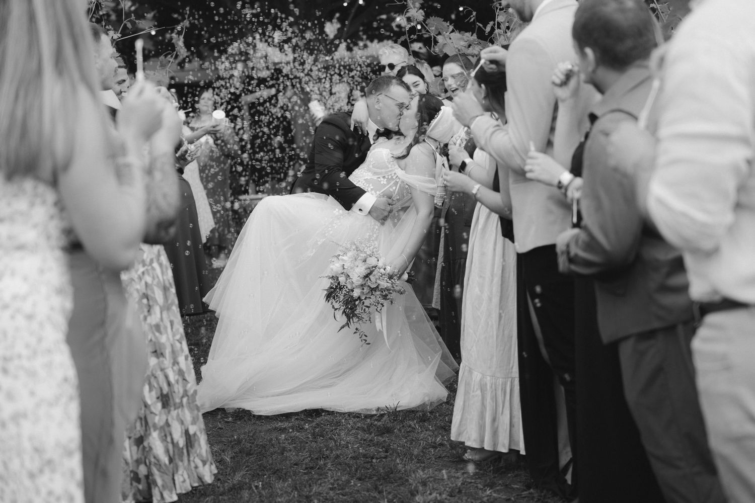 Wedding guests celebrate and toss flower petals at a joyful outdoor garden ceremony in black and white.