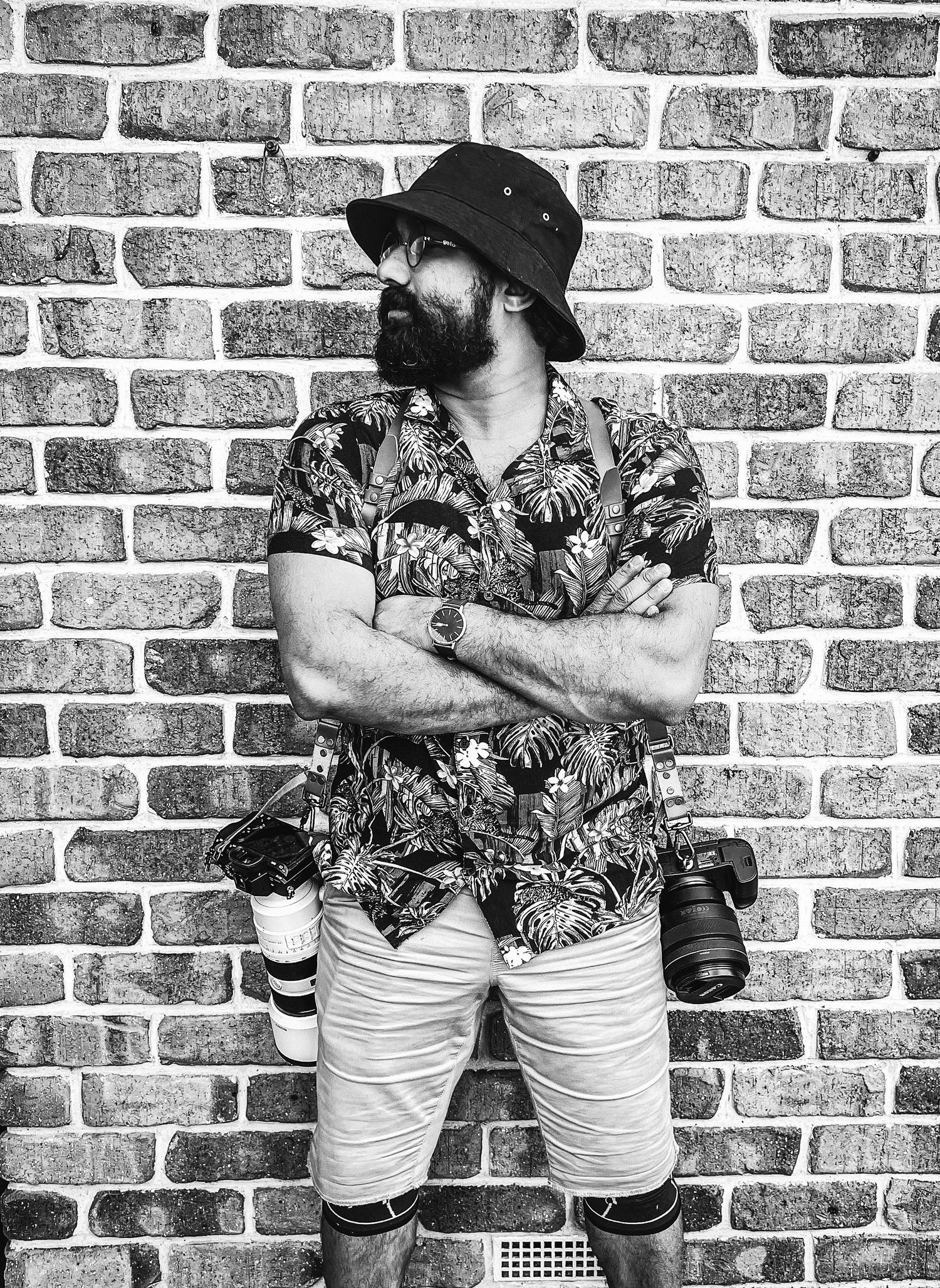 Black and white portrait of someone in a bucket hat and floral shirt standing against a brick wall with cameras.