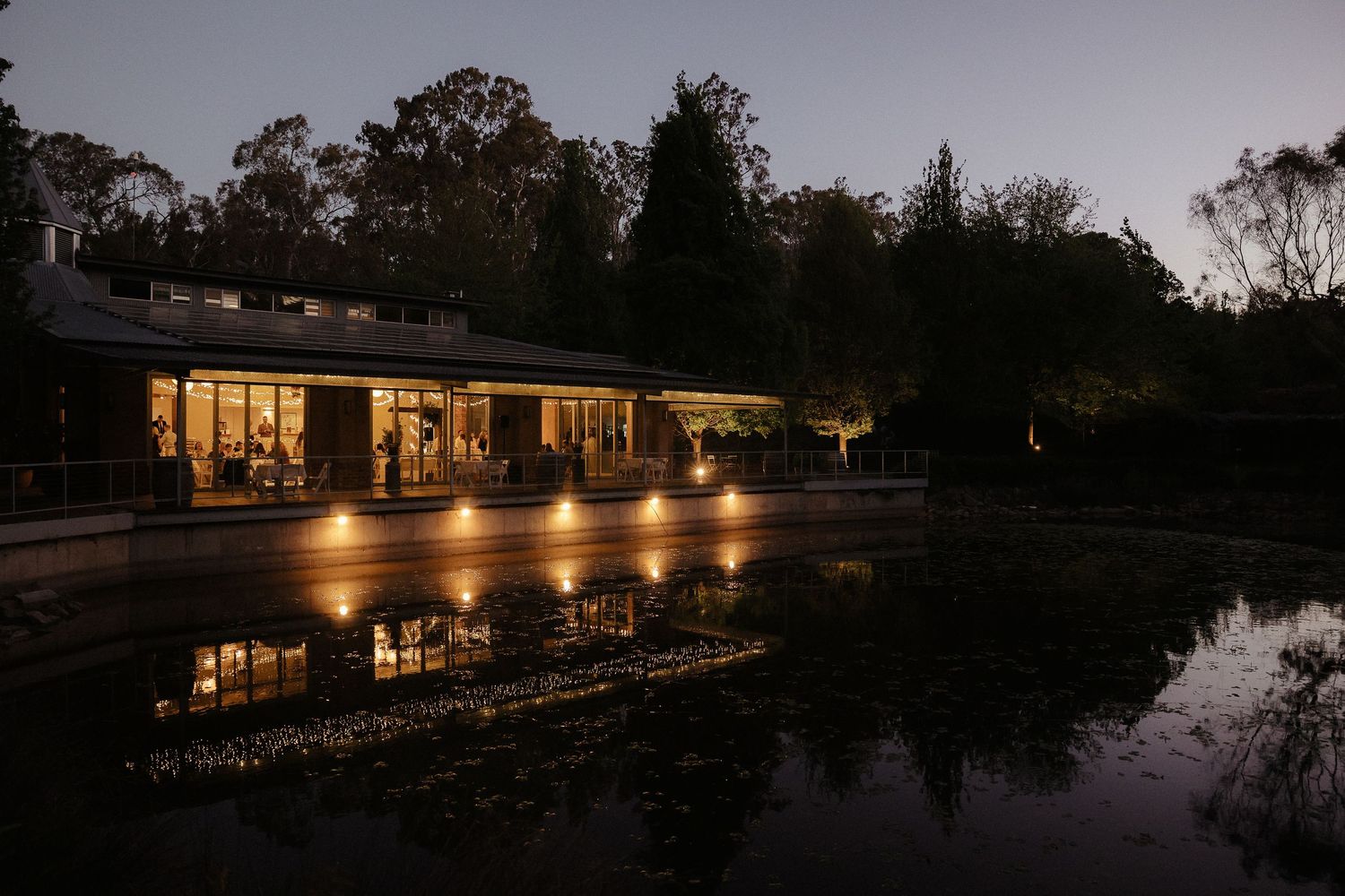 Evening exterior view of a waterfront venue with warm lighting reflecting on the water.