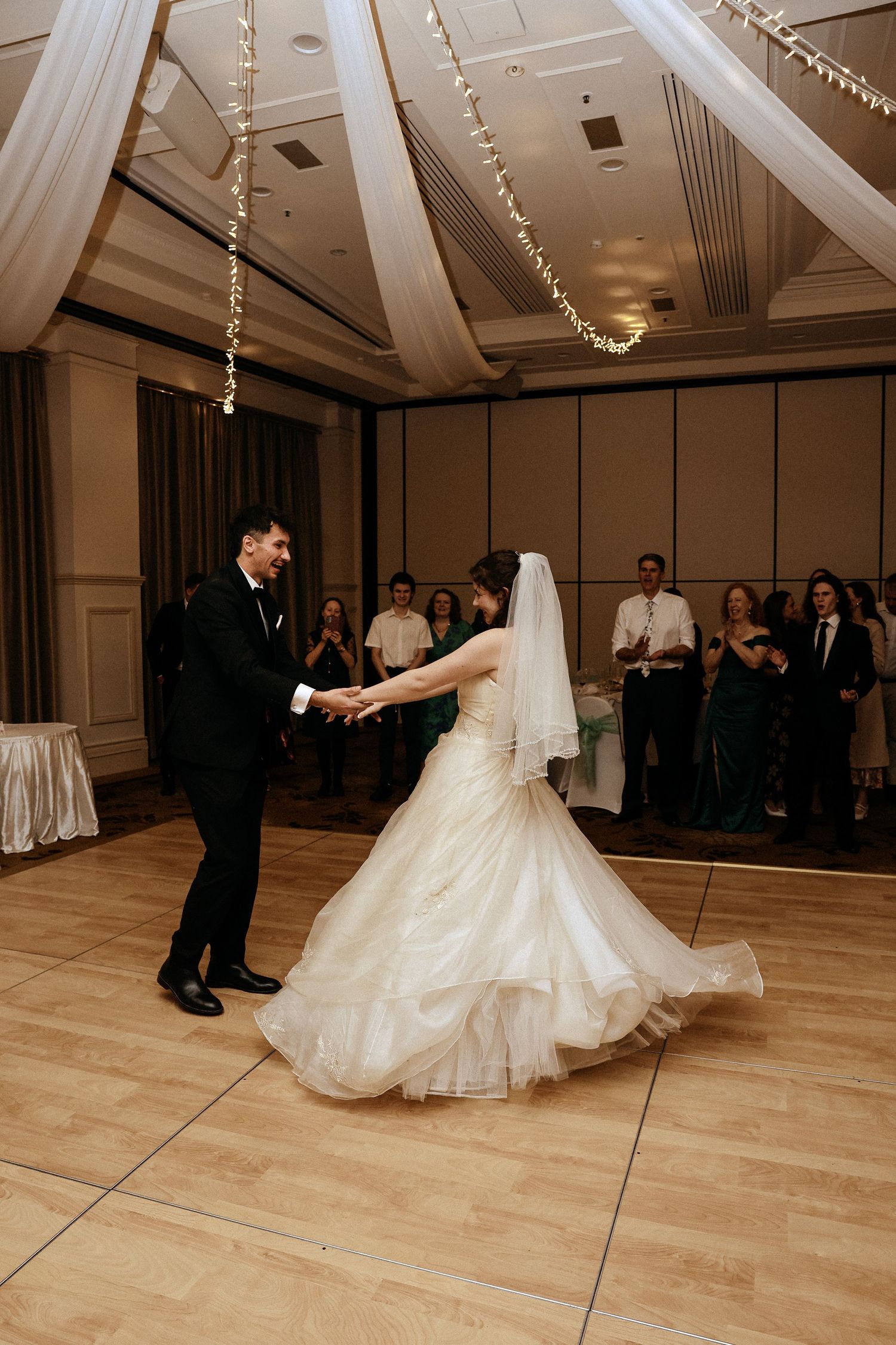 A couple performs their first dance at a wedding reception in an elegant ballroom with hardwood floors and string lights.