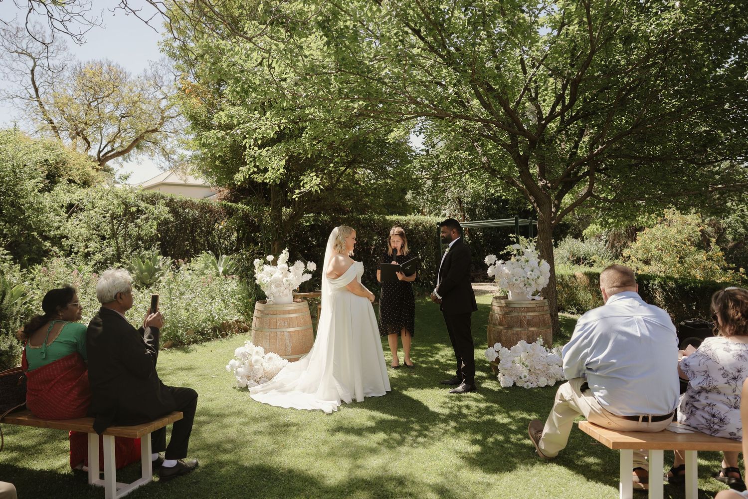 An intimate outdoor garden wedding ceremony takes place under shady trees with guests seated on wooden chairs.