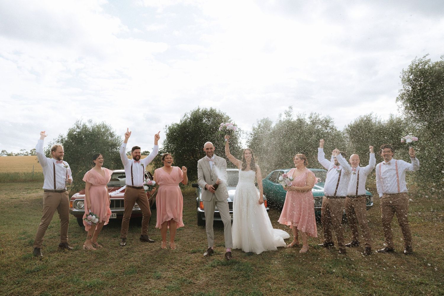 Wedding party in pink and white attire jumps joyfully together in a garden setting during sunset.