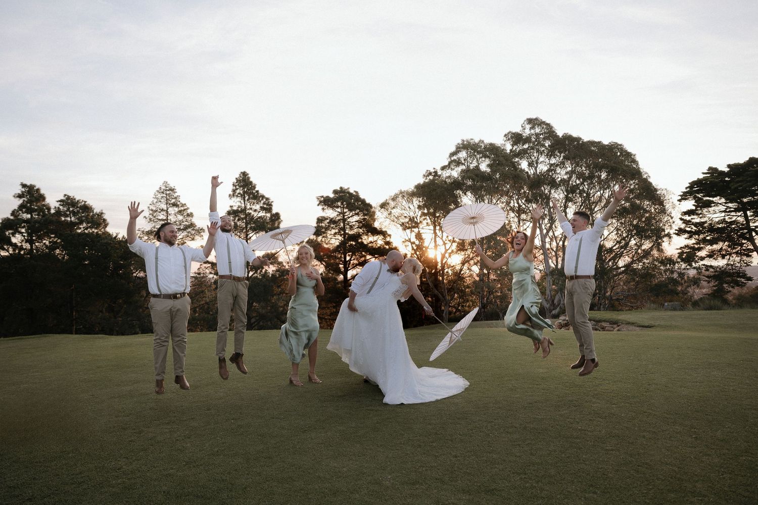 Wedding party jumps joyfully on a grassy field at sunset with trees silhouetted in the background.