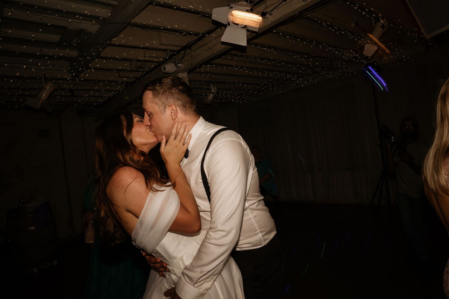 A couple embraces while dancing at an indoor celebration with dark ambient lighting.