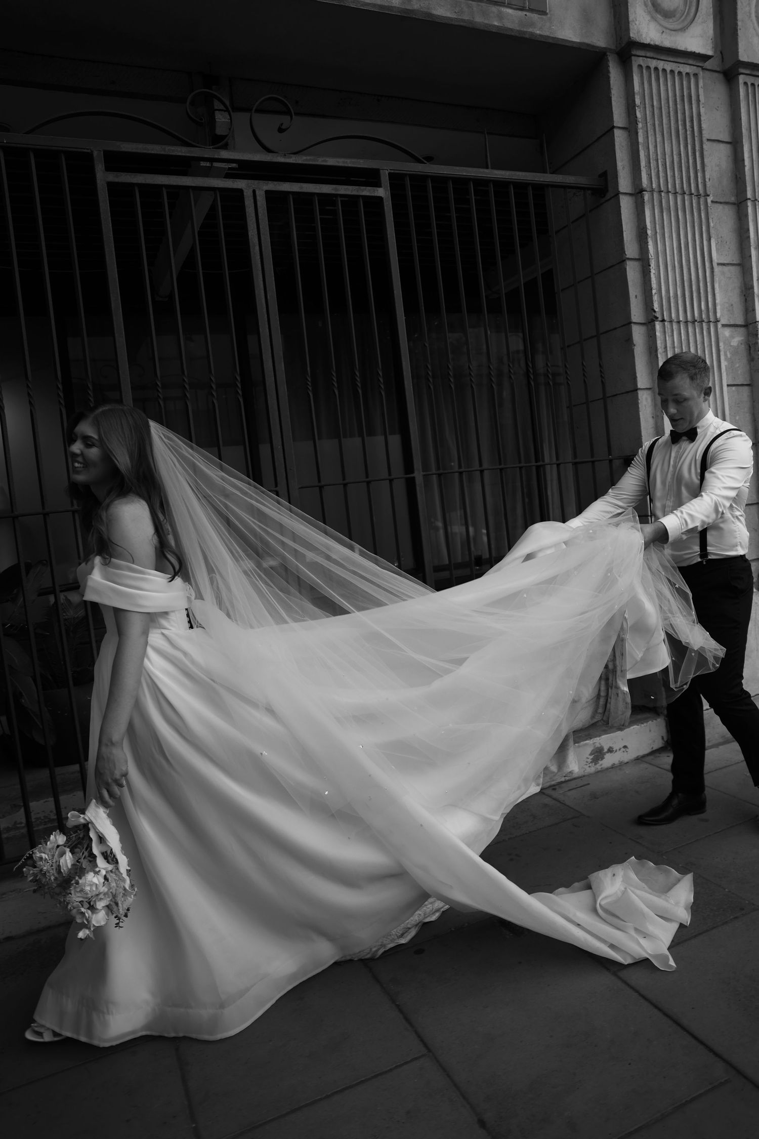 A dramatic black and white photo of a wedding dress with a flowing long train being carried on a city street.