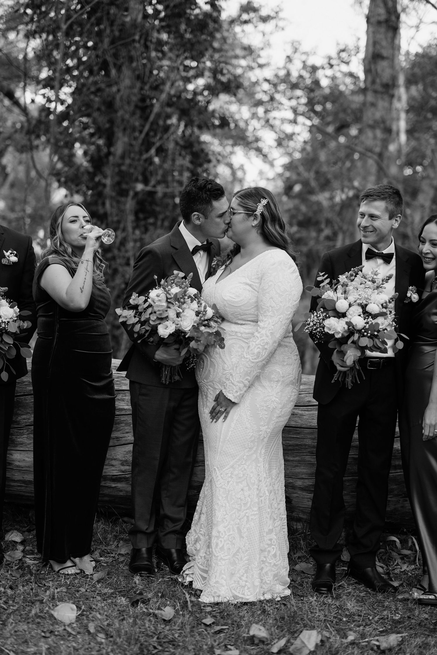 Wedding party in black attire stands in line outdoors while newlyweds share romantic moments in white dress.