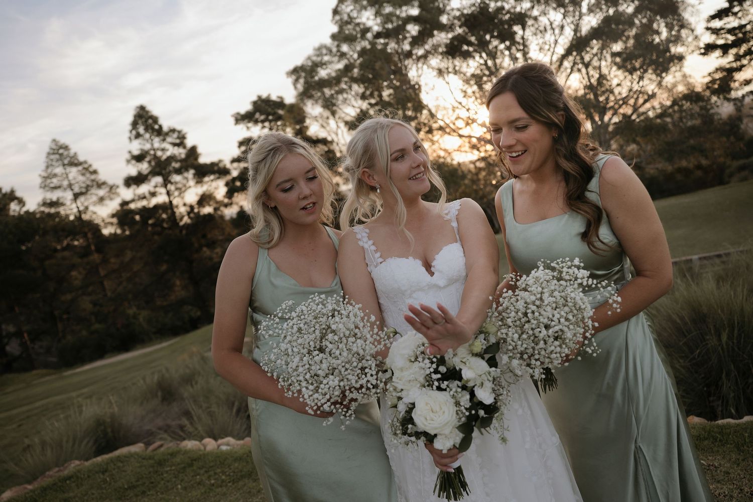 Three people in formal attire hold white baby's breath bouquets while sharing a joyful moment at an outdoor wedding.