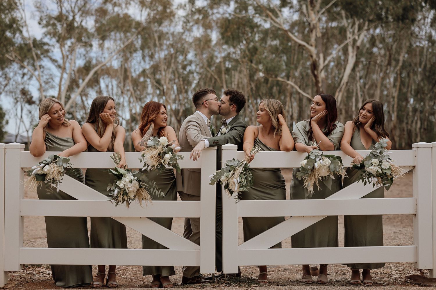 A romantic outdoor wedding celebration with couples sitting at a rustic white fence during golden hour.