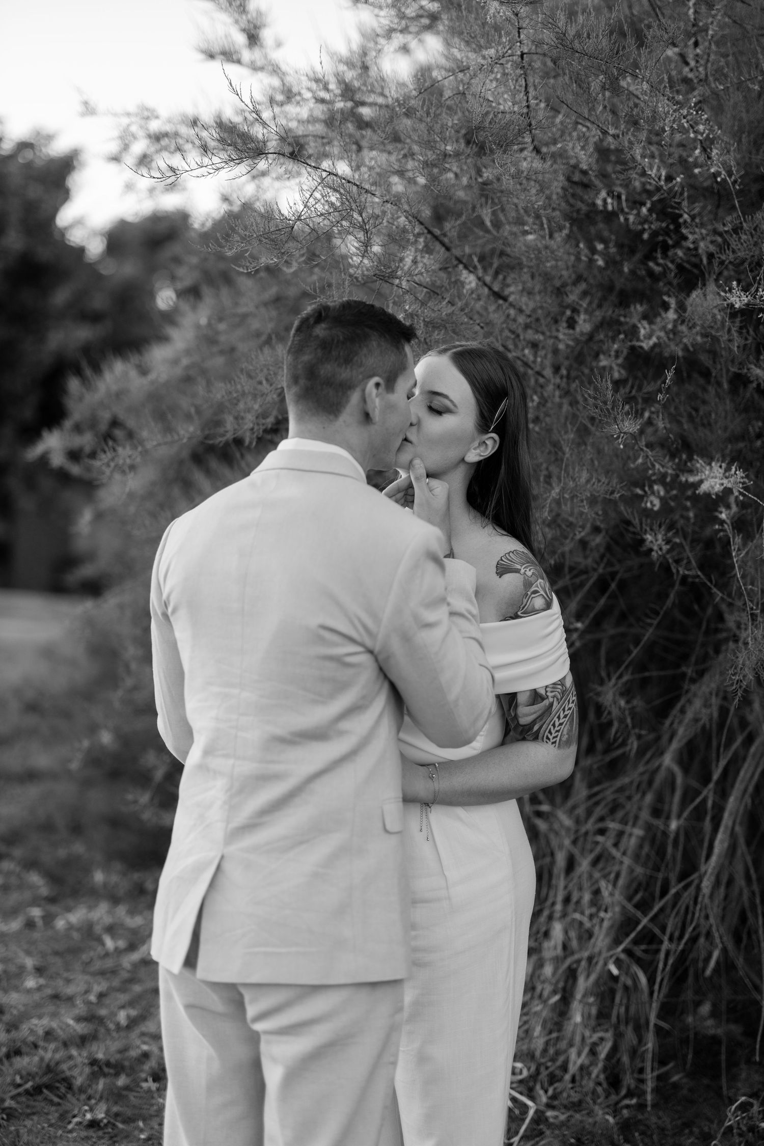 A romantic black and white photograph of a couple sharing a tender kiss outdoors among natural foliage.