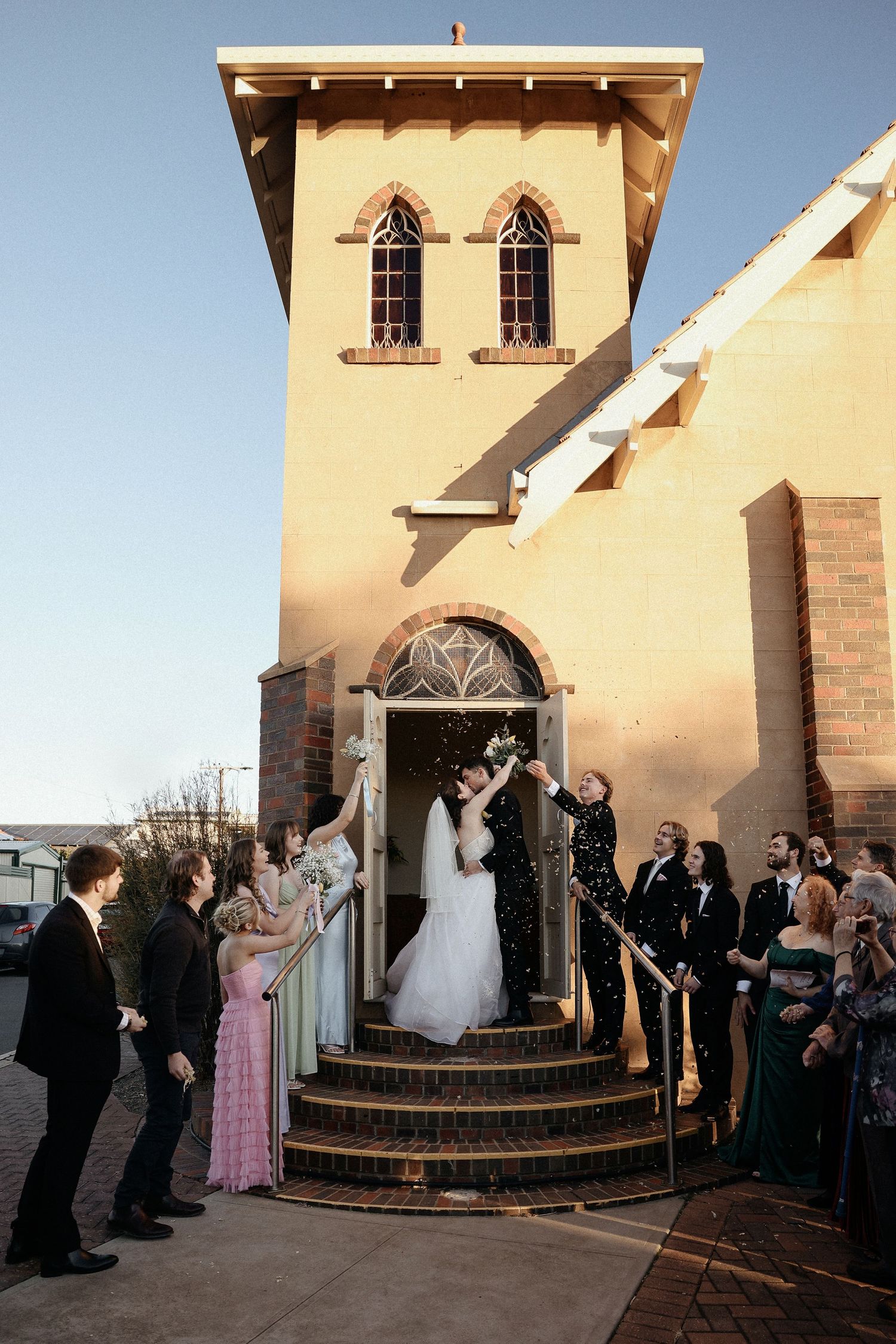 Wedding ceremony taking place at sunset outside an adobe-style church with guests gathered around curved steps.