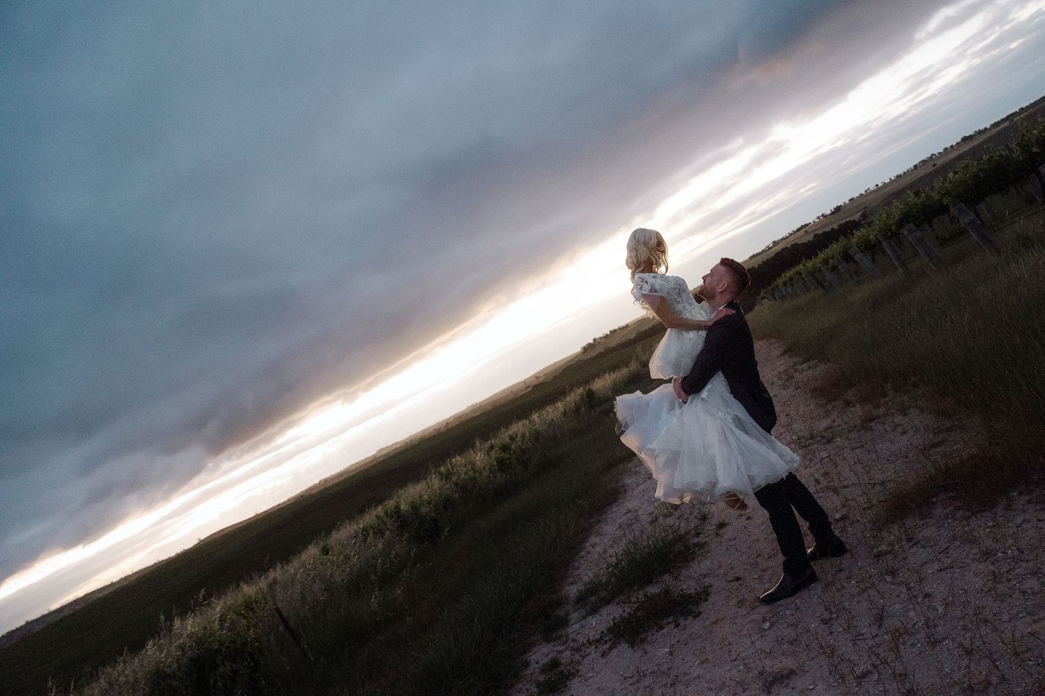 A couple in wedding attire embraces on a hillside at sunset with dramatic stormy clouds overhead.