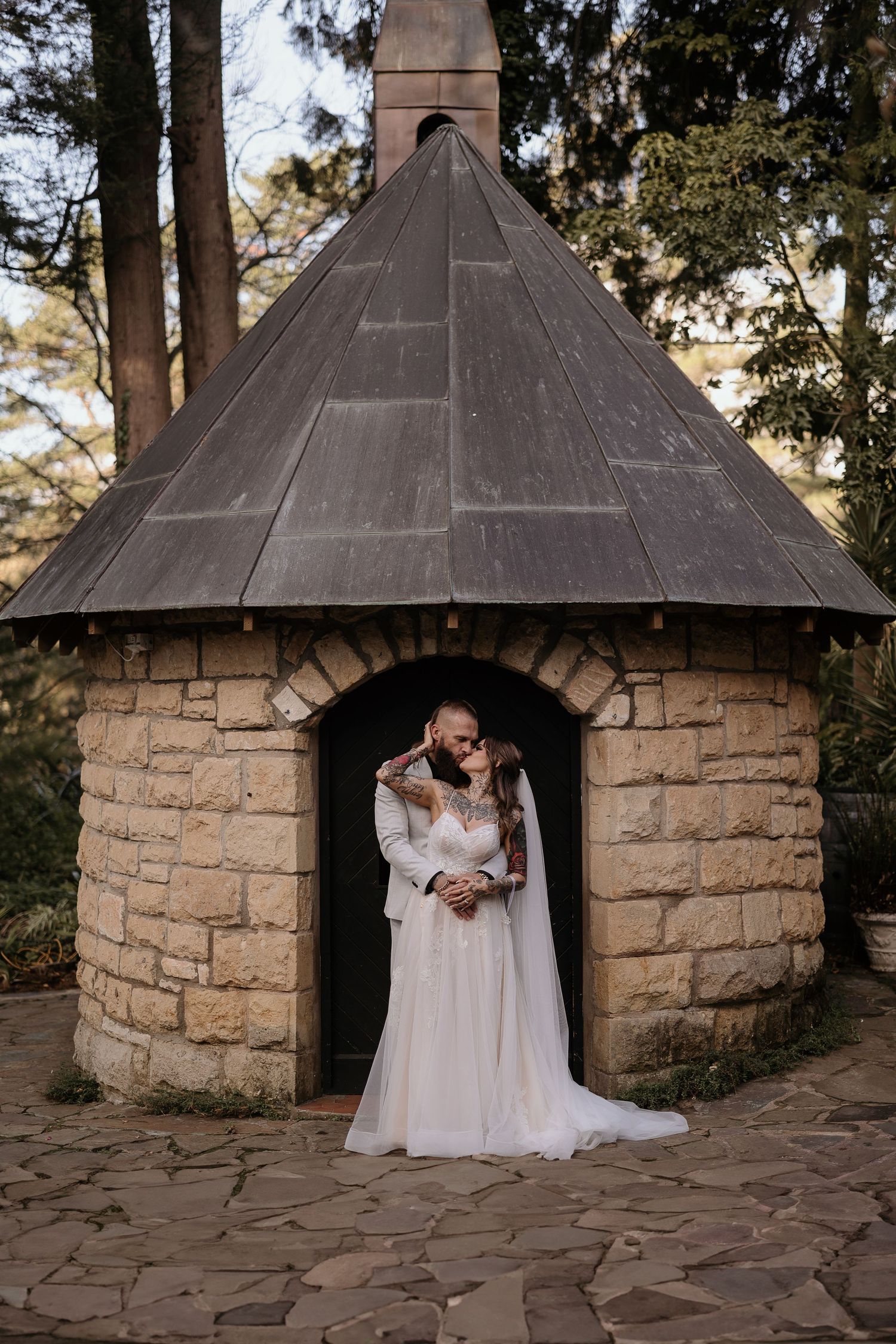A stone gazebo with conical roof stands among tall trees in a rustic outdoor wedding setting.