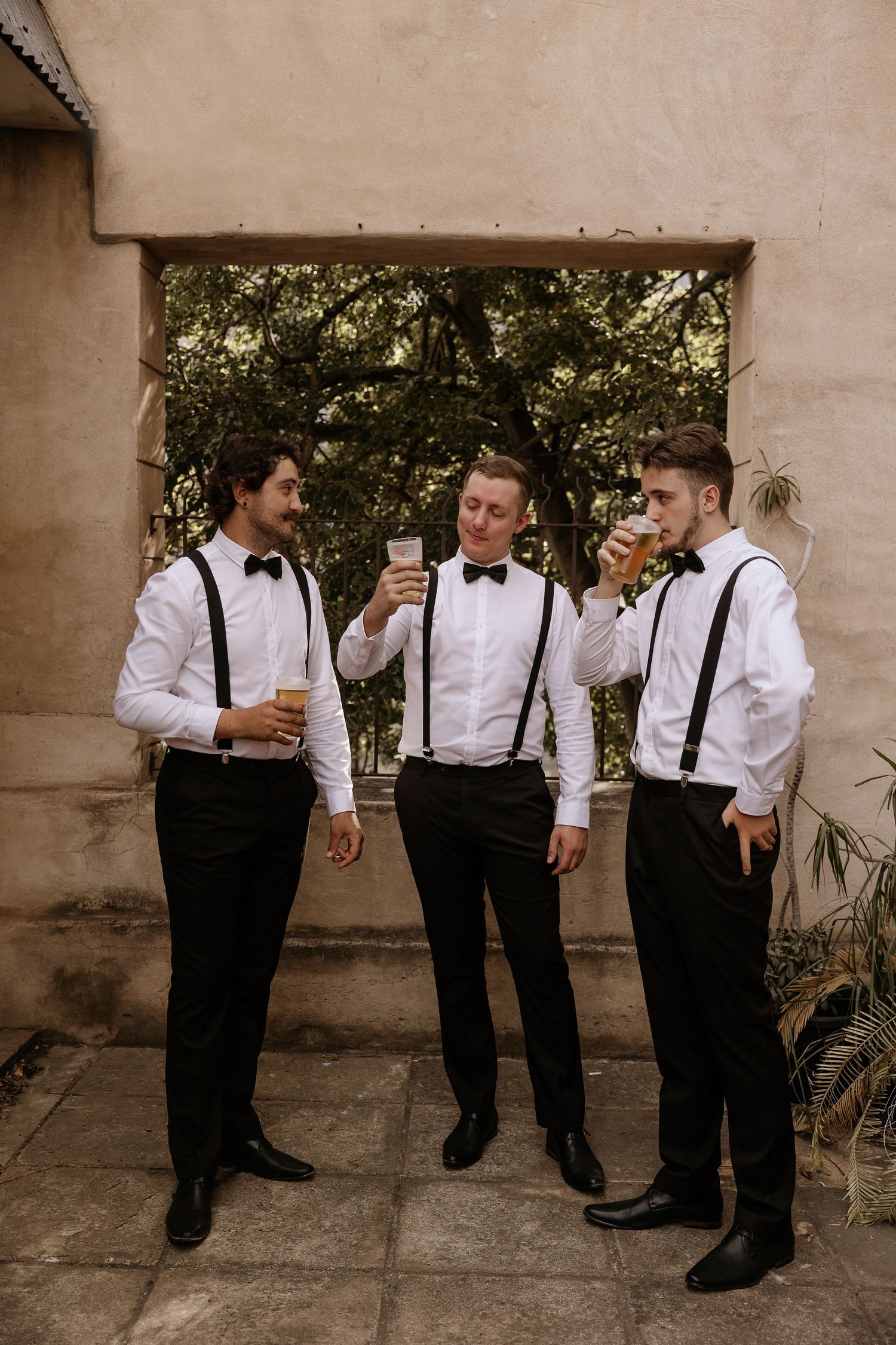 Groomsmen in white shirts and suspenders share drinks and laughs during wedding preparations.