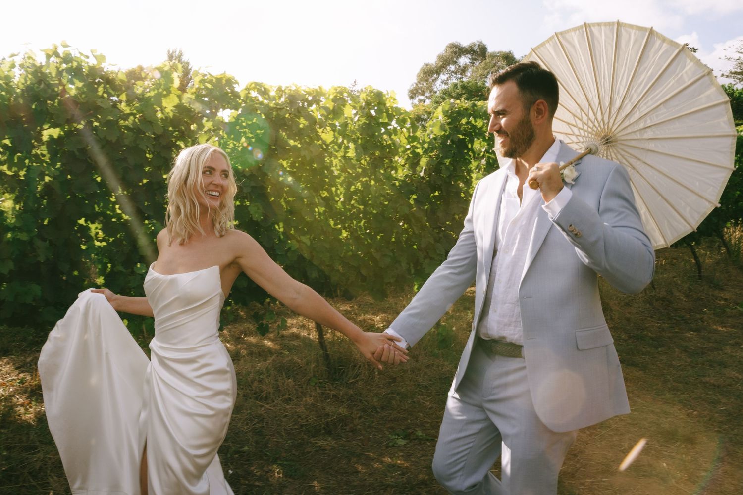 Wedding couple poses with white parasol in golden sunset light among garden foliage.