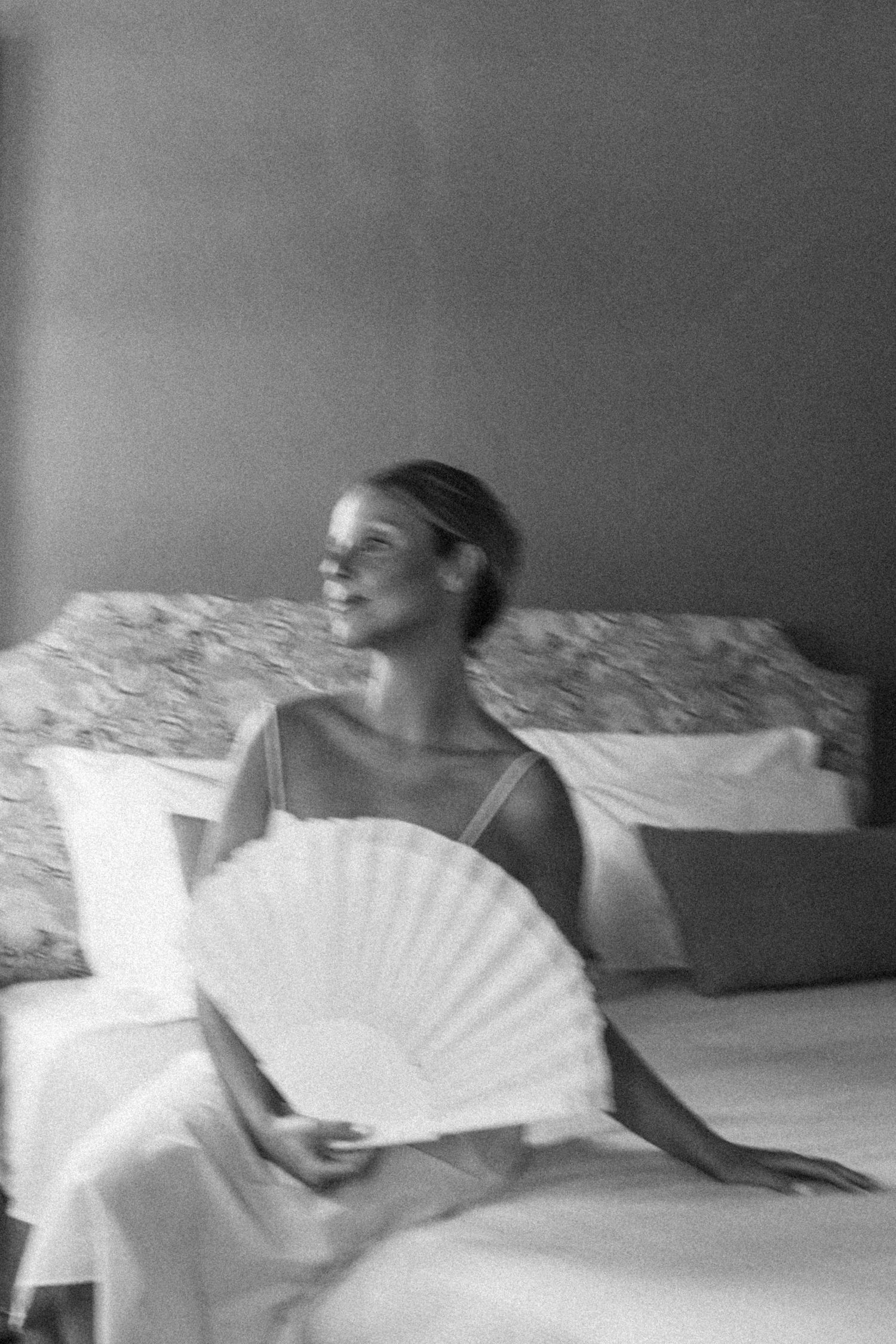 An elegant scene of someone holding a white paper fan while sitting on a bed in black and white.