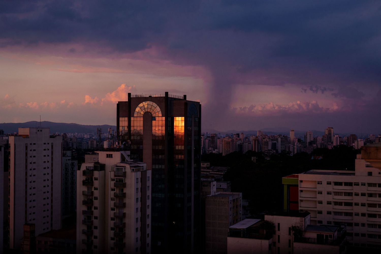 Urban skyline silhouette at sunset with buildings backlit against a purple and orange sky.