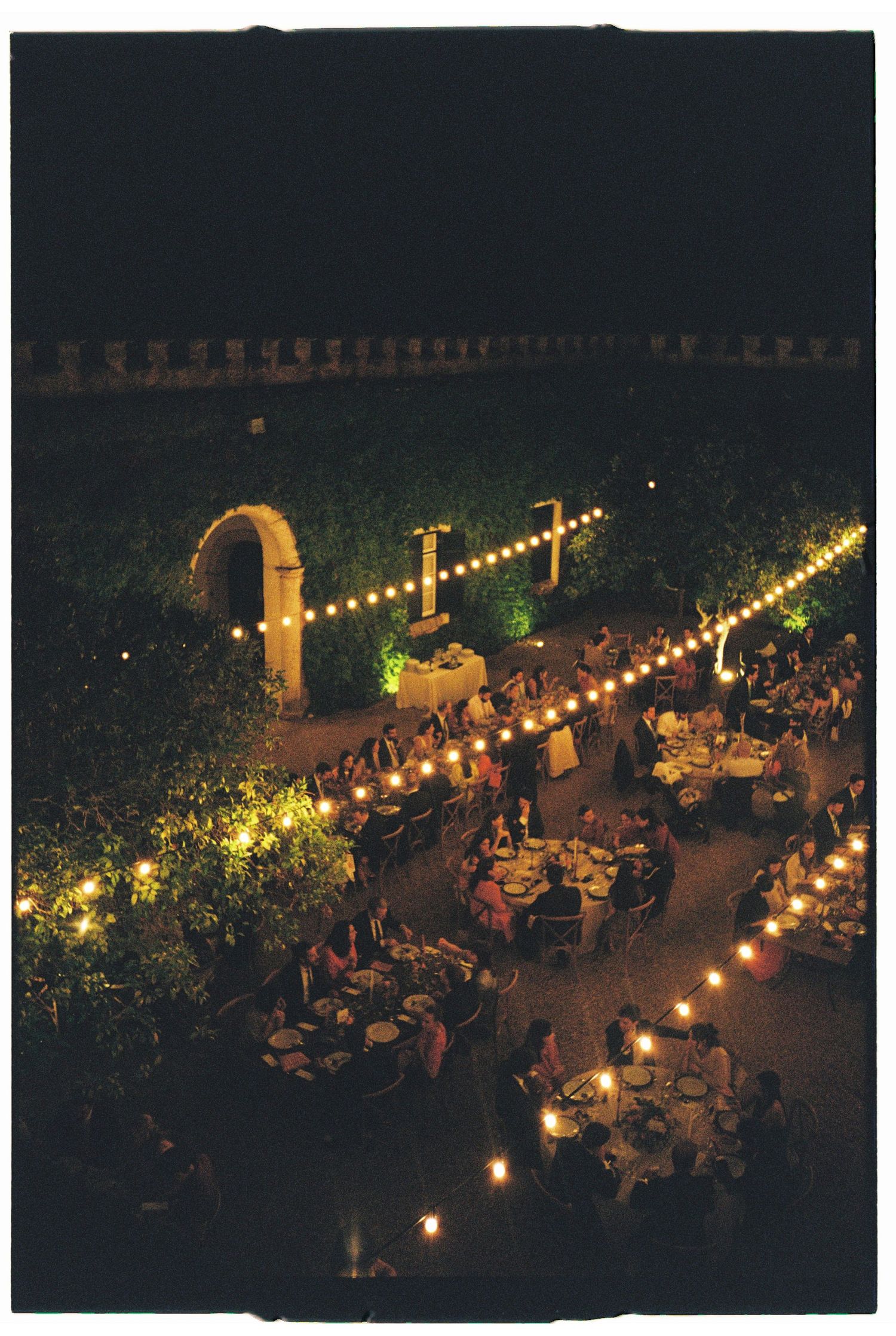 Outdoor evening dinner reception with string lights illuminating tables and guests under a romantic archway.