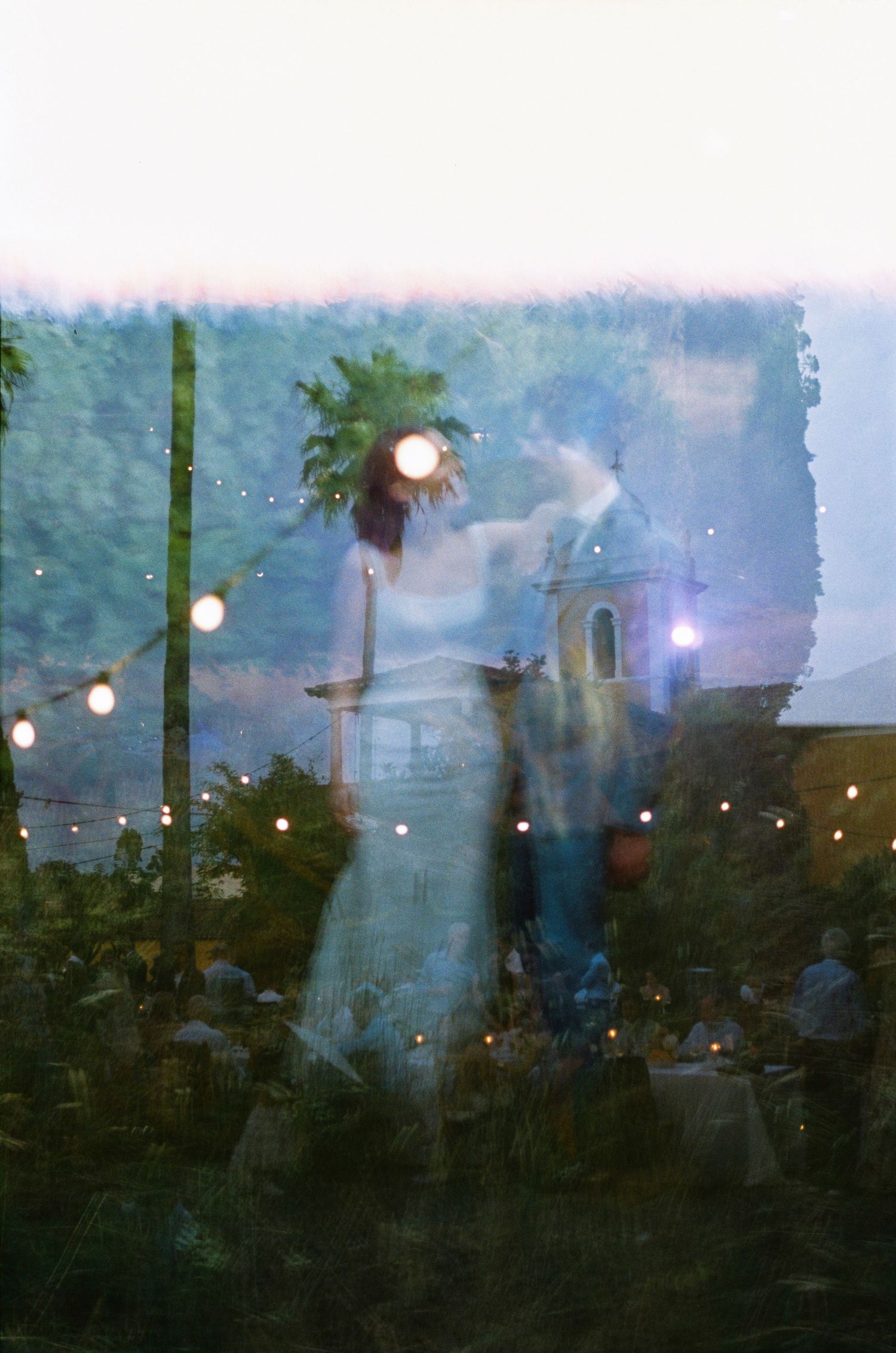 Motion blur captures people dancing and celebrating at an outdoor event at dusk with glowing lights in the background.